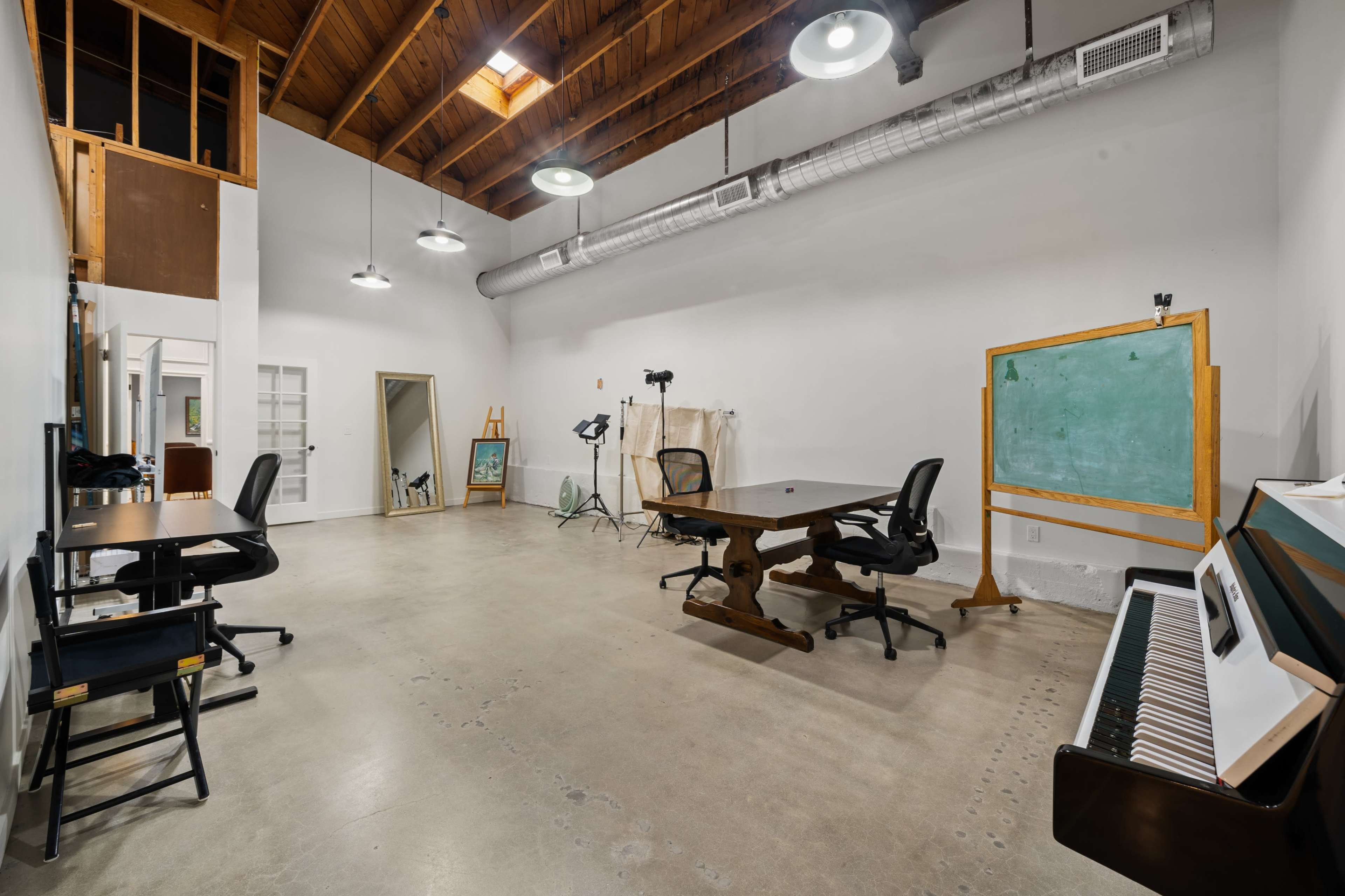 The image shows a spacious, well-lit studio area featuring a large table, several chairs, a chalkboard, and a piano, with a high wooden ceiling and industrial lighting fixtures.