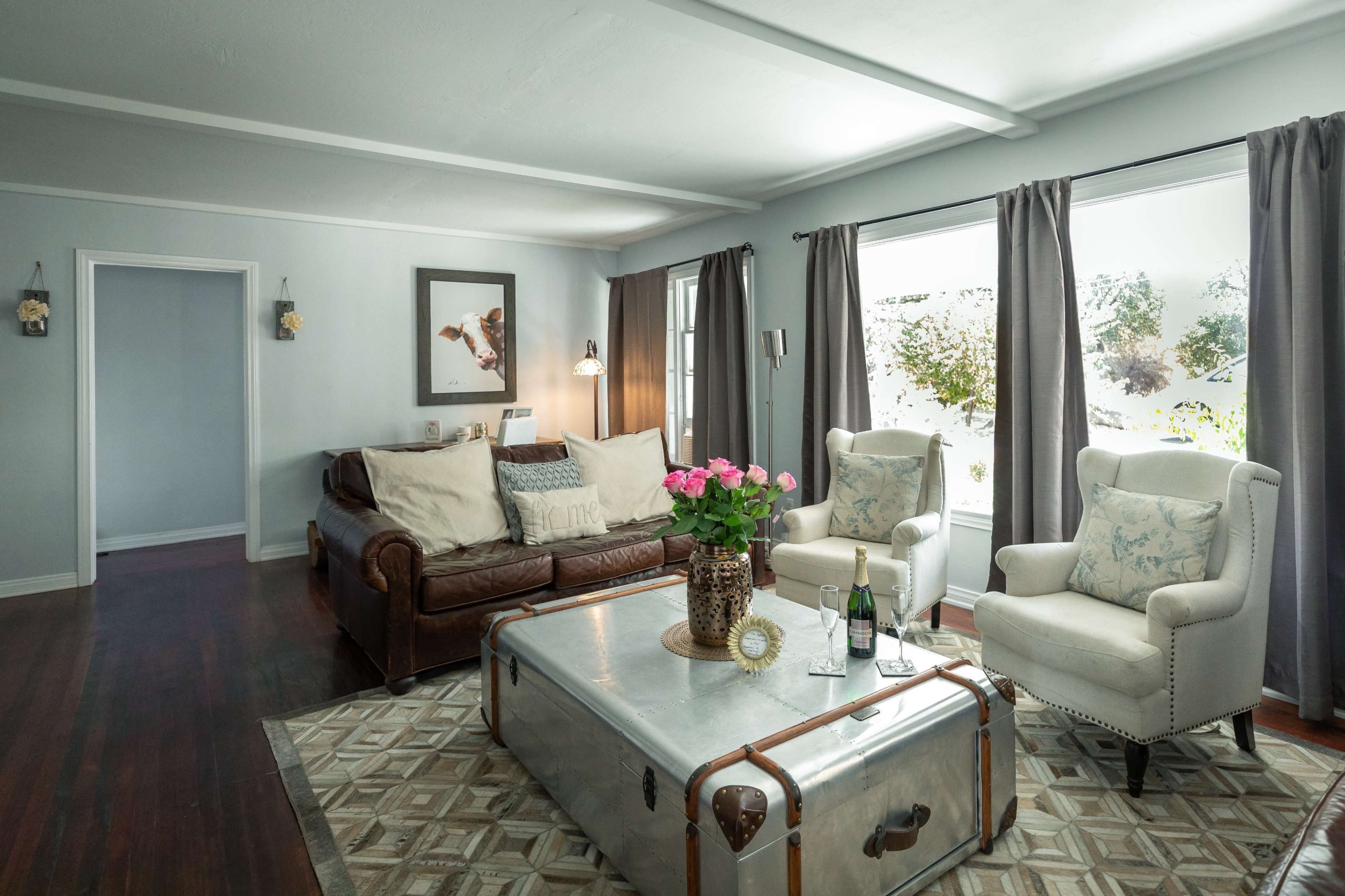 A living room with a brown leather sofa, two white armchairs, a coffee table, and a window with natural light illuminating the space.