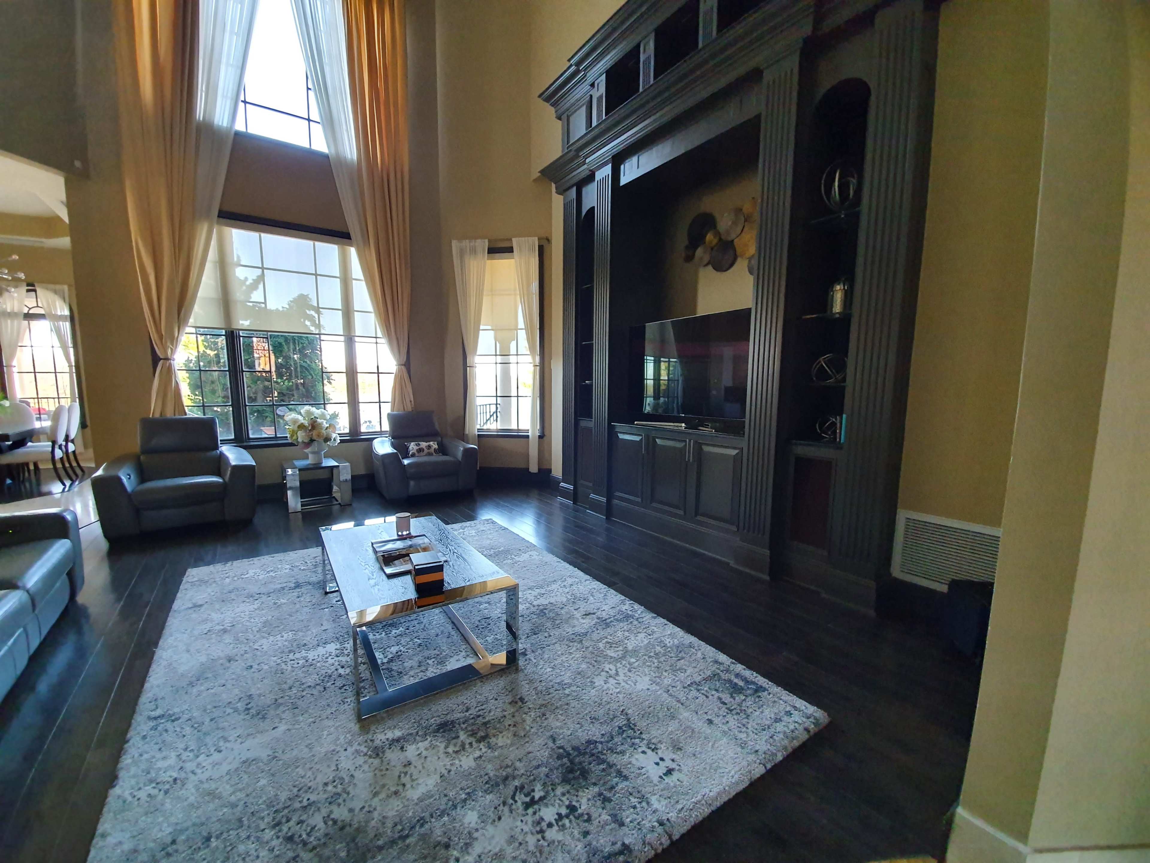 A spacious living room with large windows, a dark wooden entertainment unit, two gray armchairs, and a central coffee table on a patterned rug.