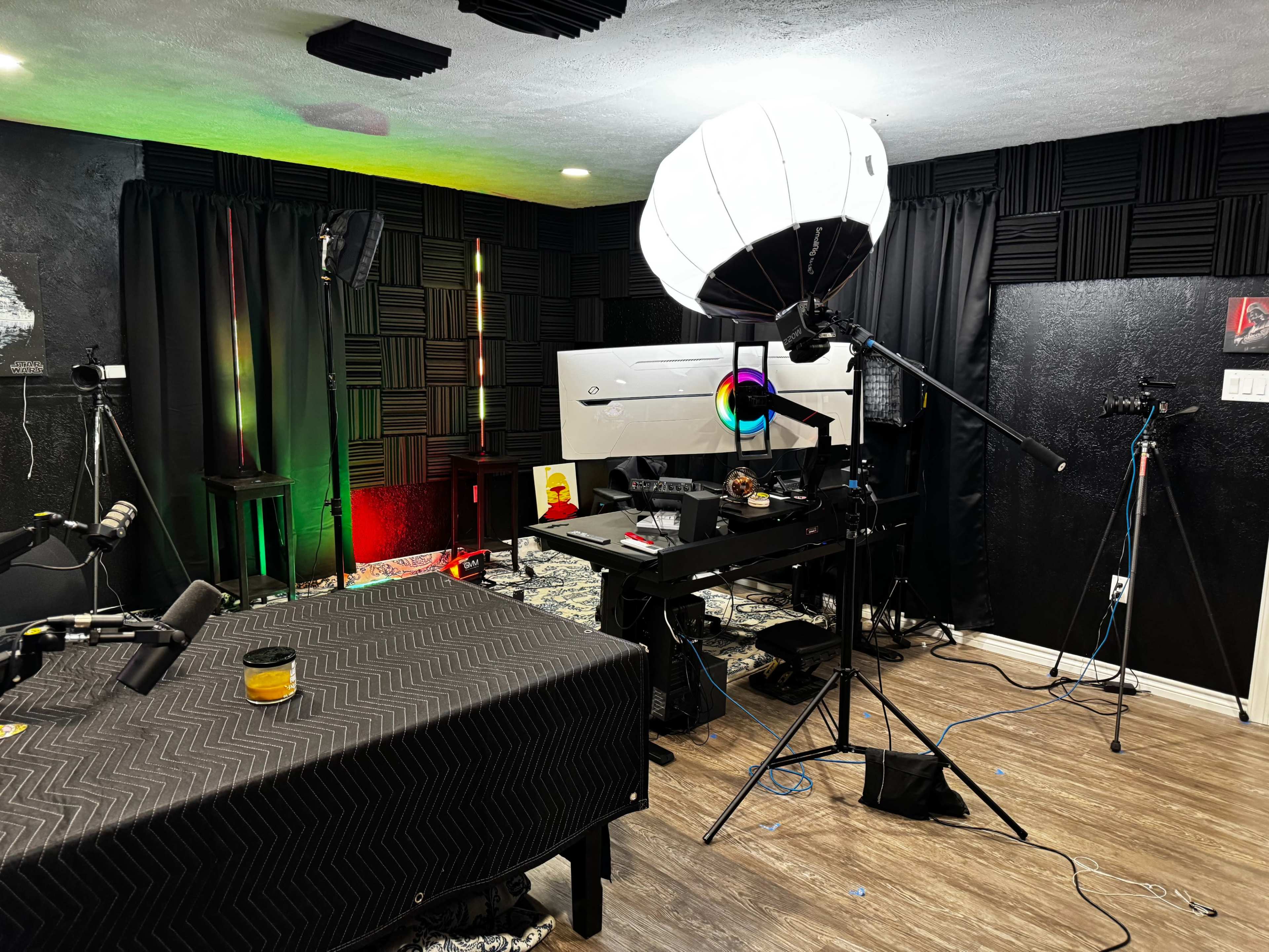 The image shows a dimly lit room set up for video production, featuring a desk with a computer, multiple cameras, microphones, and various lighting equipment.