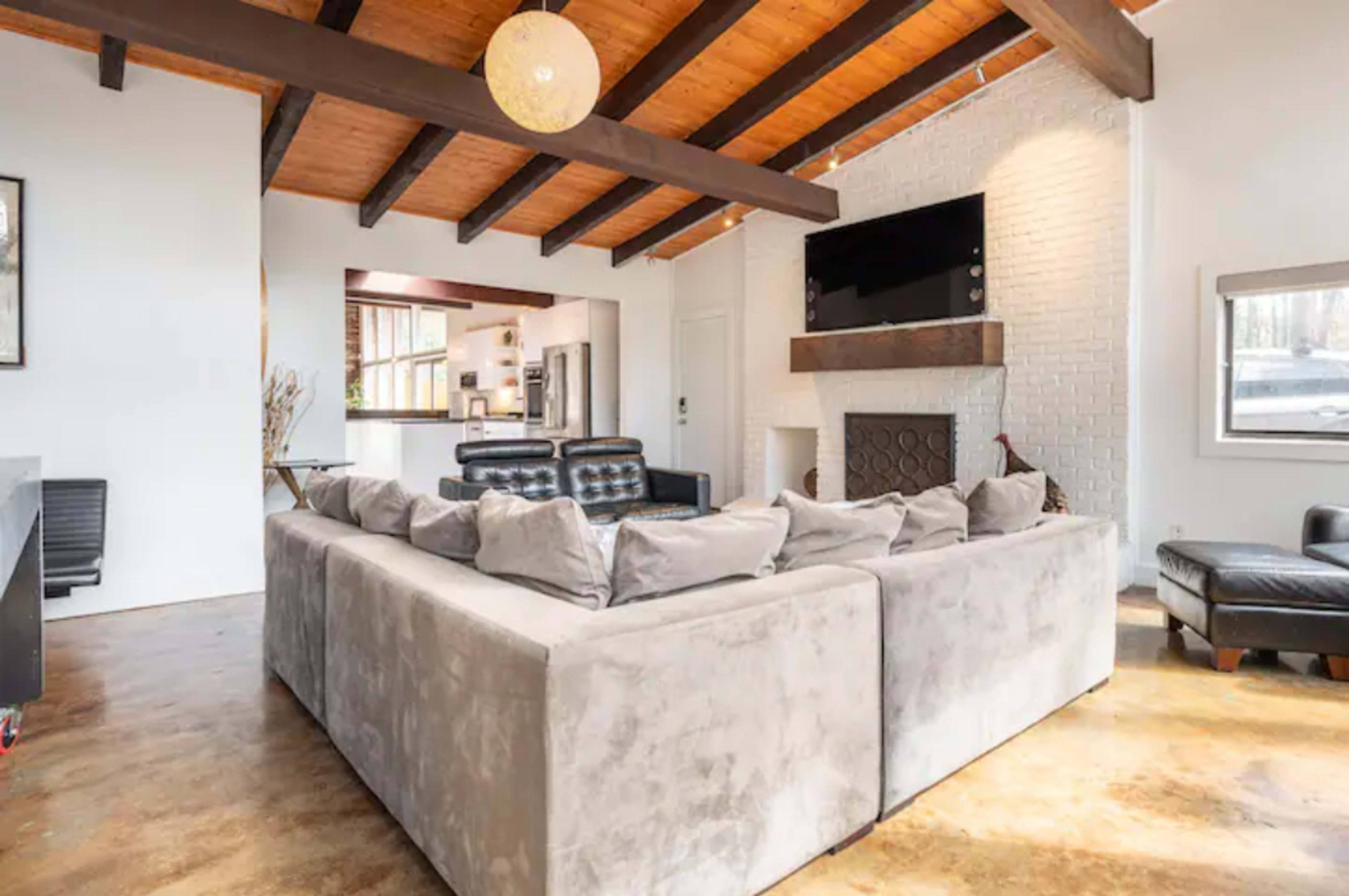 The image shows a modern living room featuring a large L-shaped sofa, wooden beams on the ceiling, and a brick fireplace with a mounted television above it.