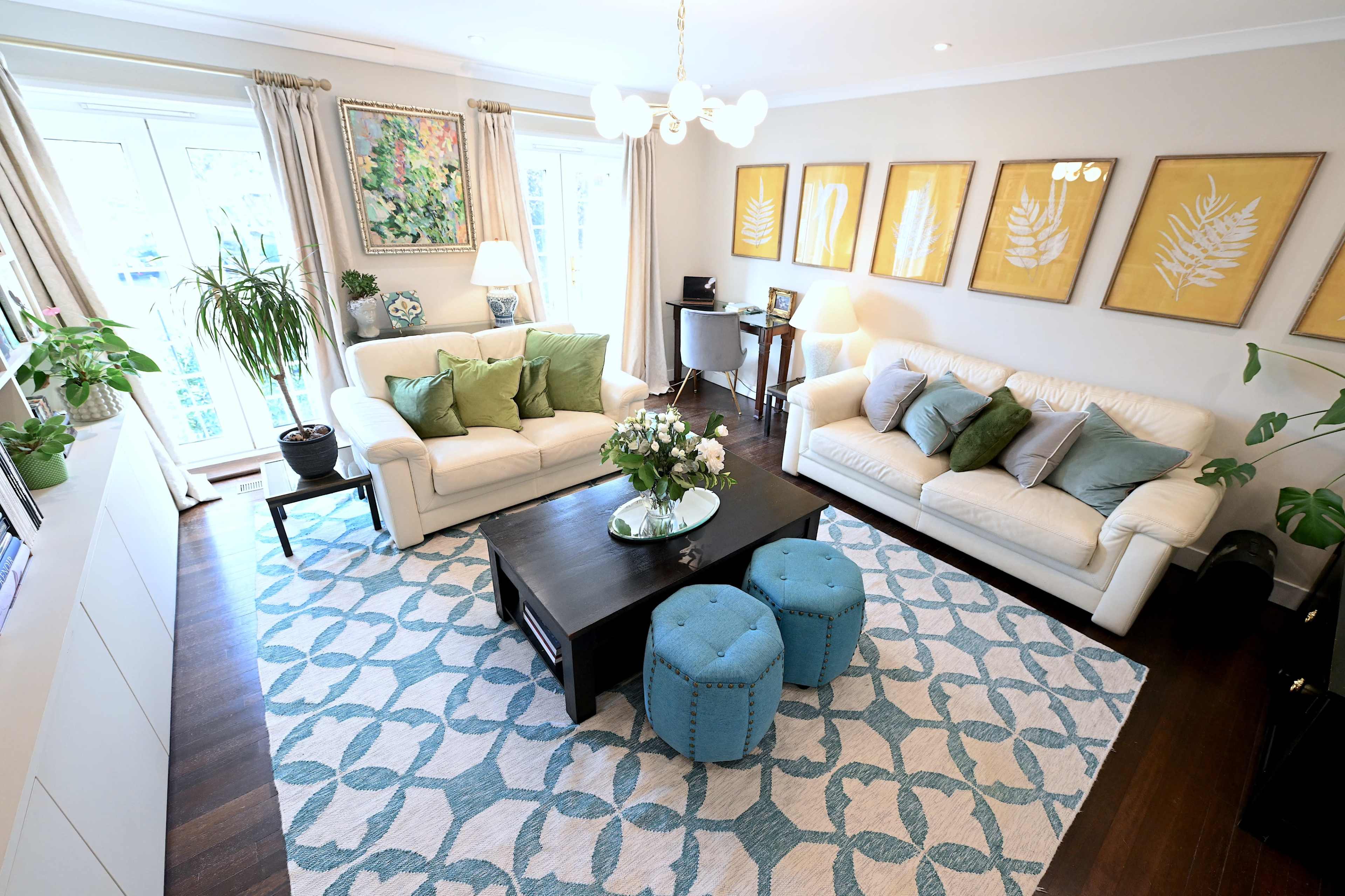 A bright living room features two white sofas, a dark coffee table, and a patterned area rug, with decorative plants and framed artwork on the walls.