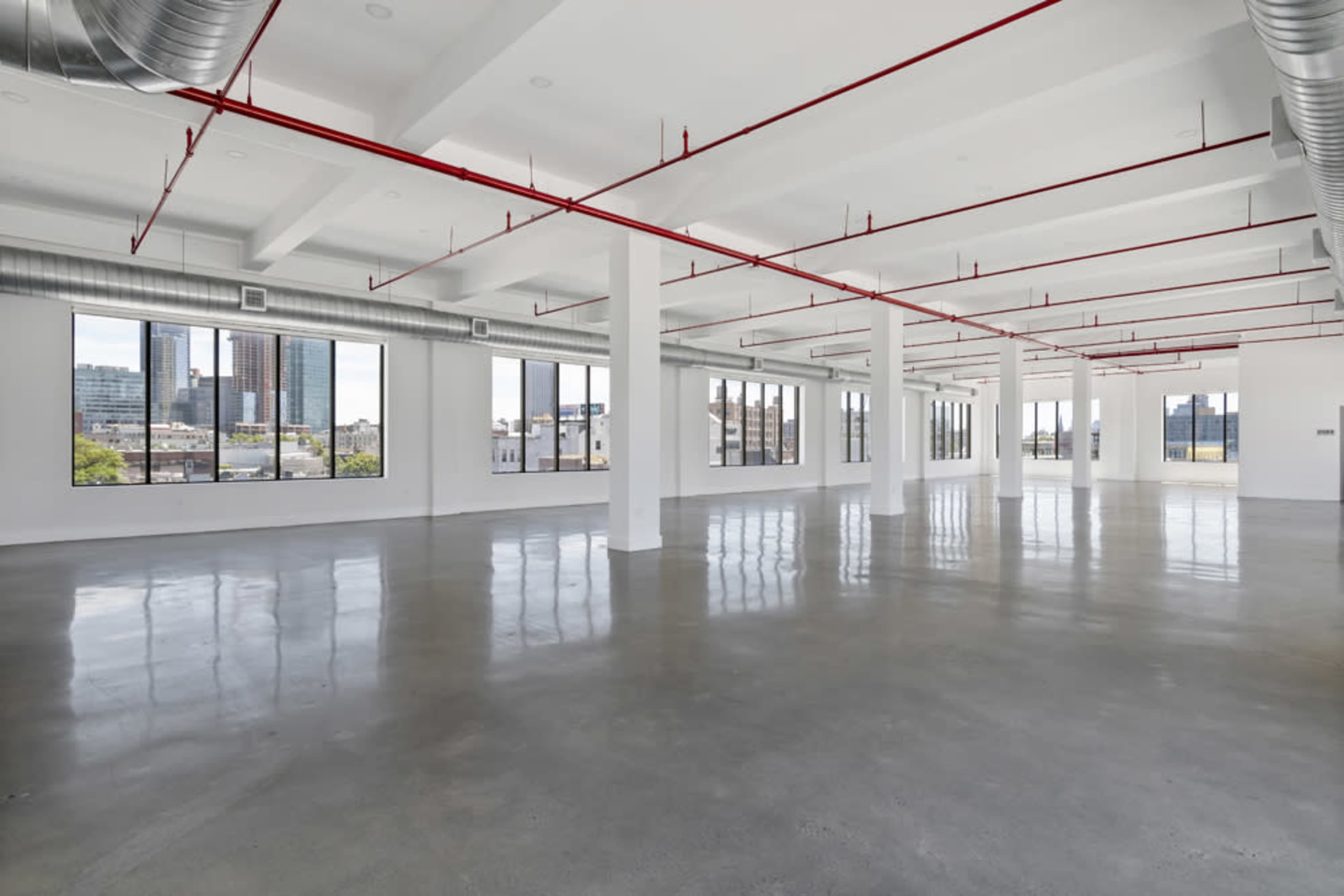 A large, empty office space with polished concrete floors and red exposed piping, featuring multiple windows that offer views of a city skyline.