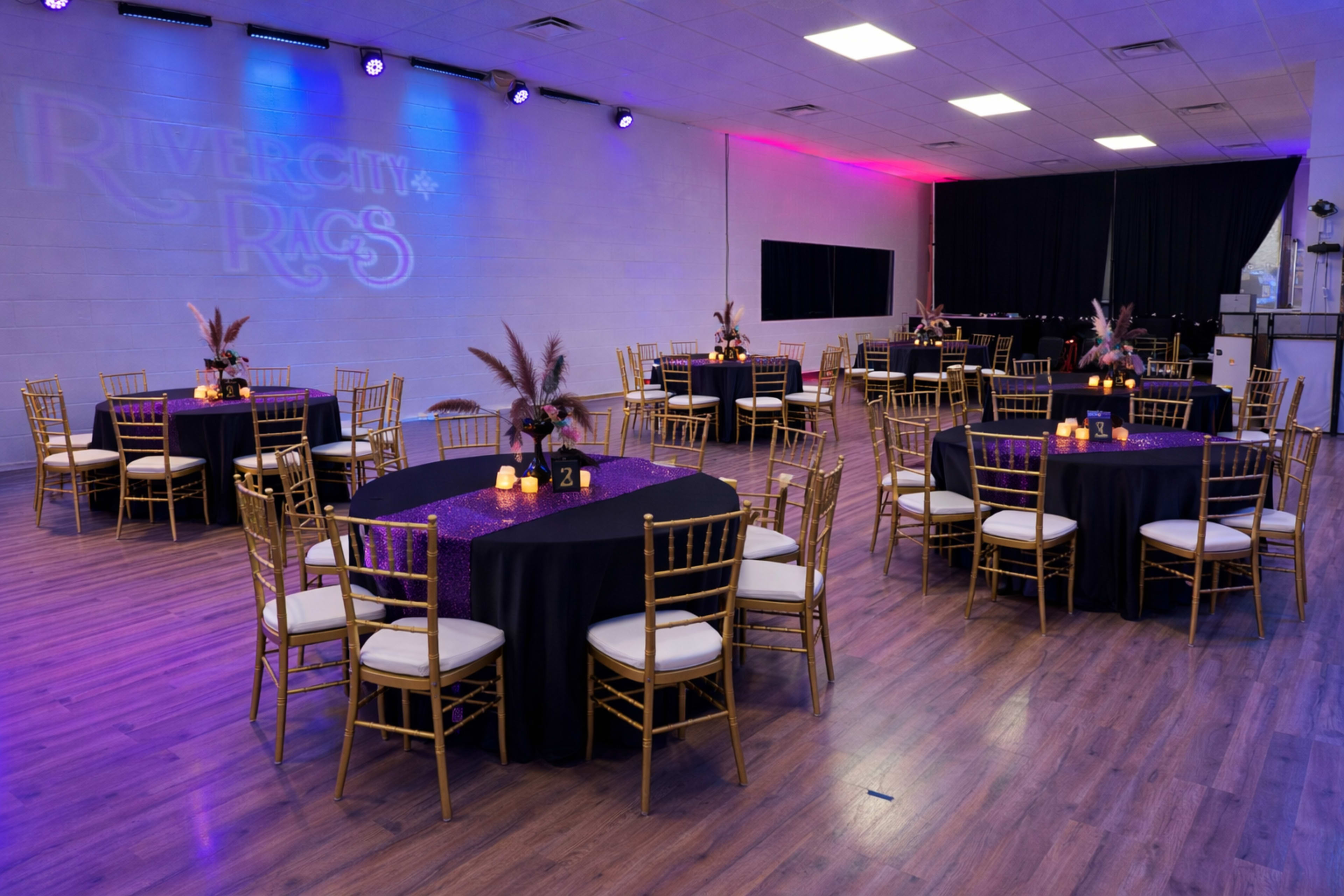 The image shows a banquet hall set up with round tables covered in black tablecloths and adorned with centerpieces, illuminated by purple and white lighting.
