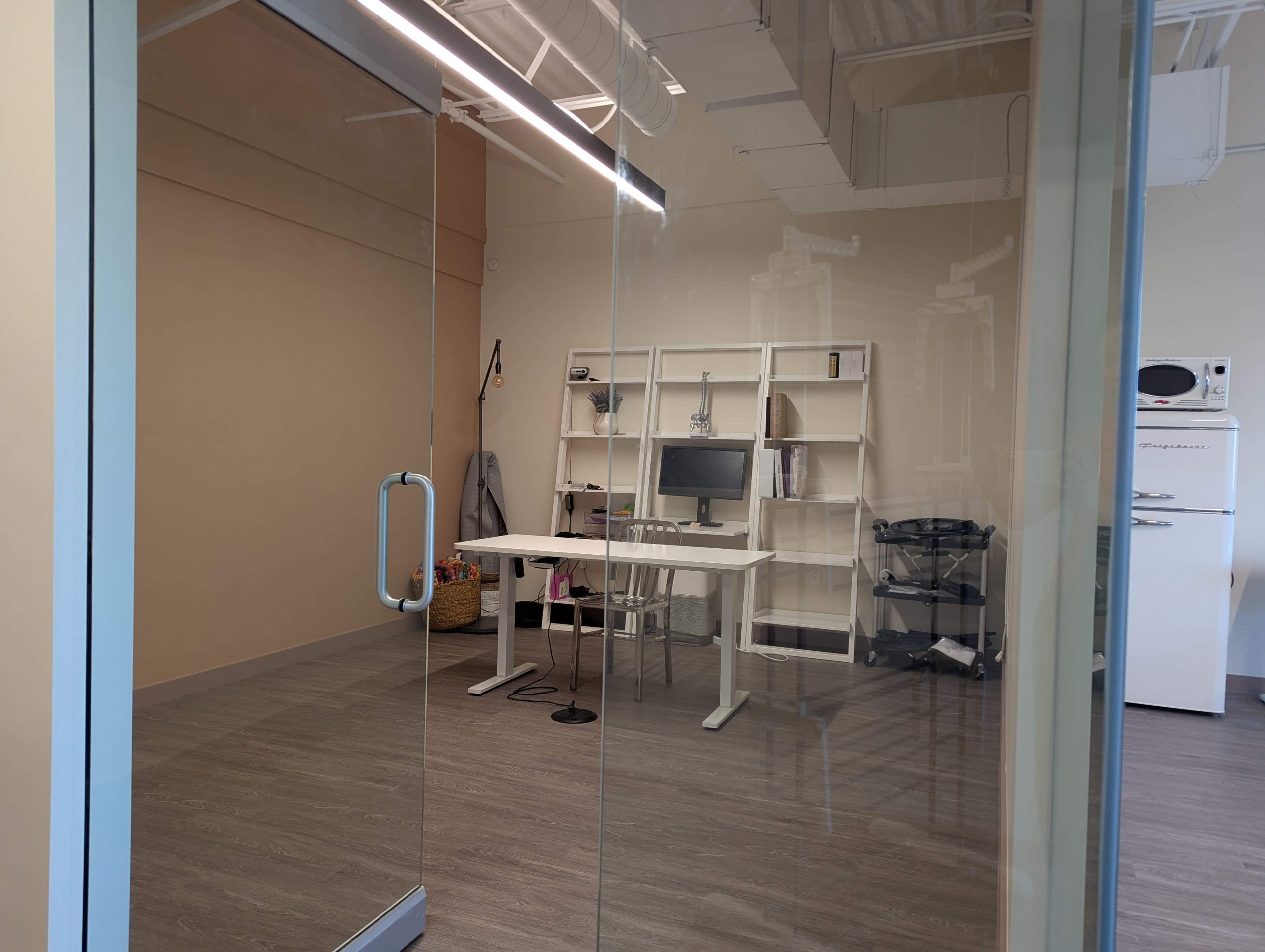 The image shows a modern office space with a glass entrance, featuring a desk with a computer, shelving, and light-colored walls.