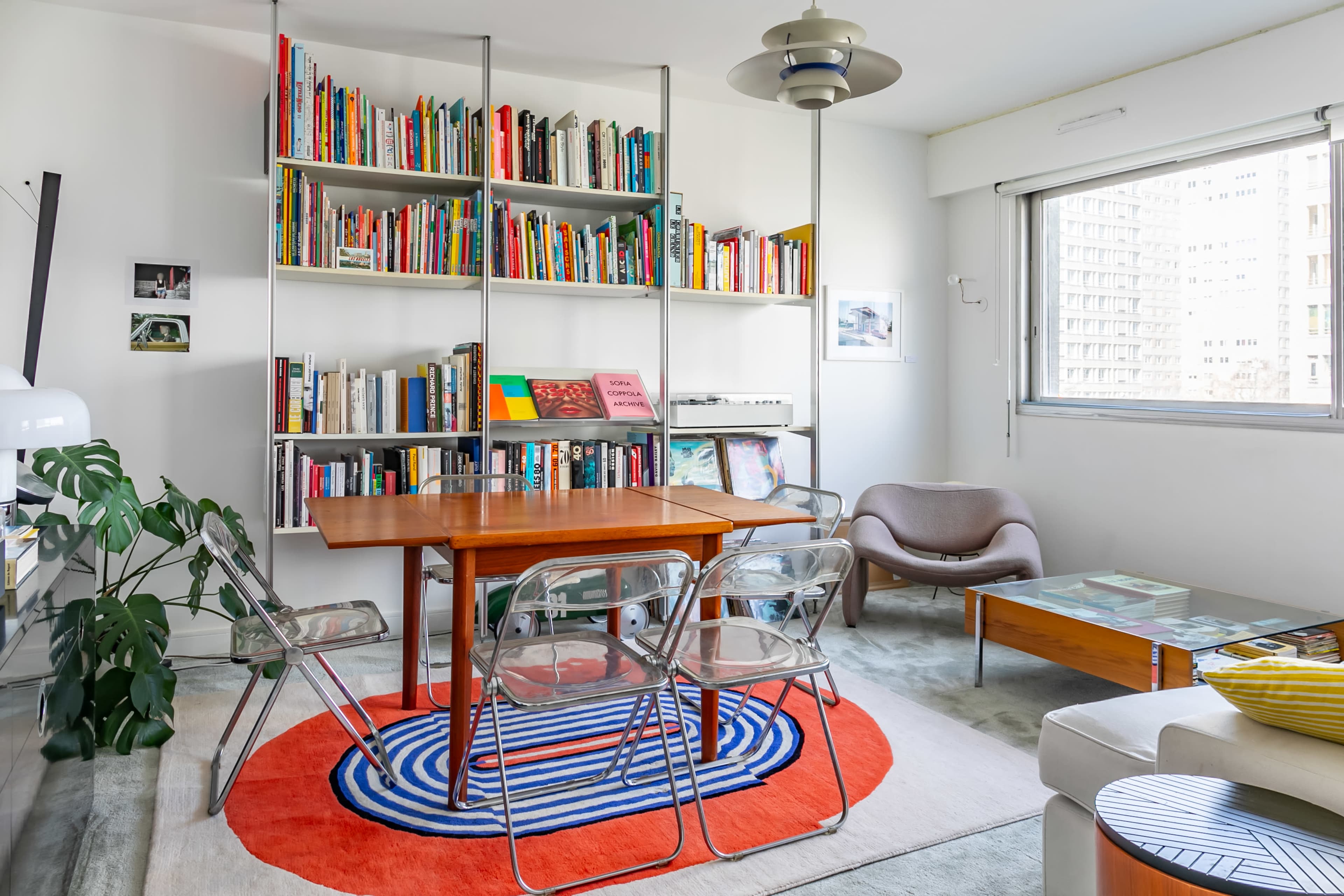 The image features a modern living space with a wooden dining table surrounded by clear chairs, a bookshelf filled with books, and a large window allowing natural light to enter.