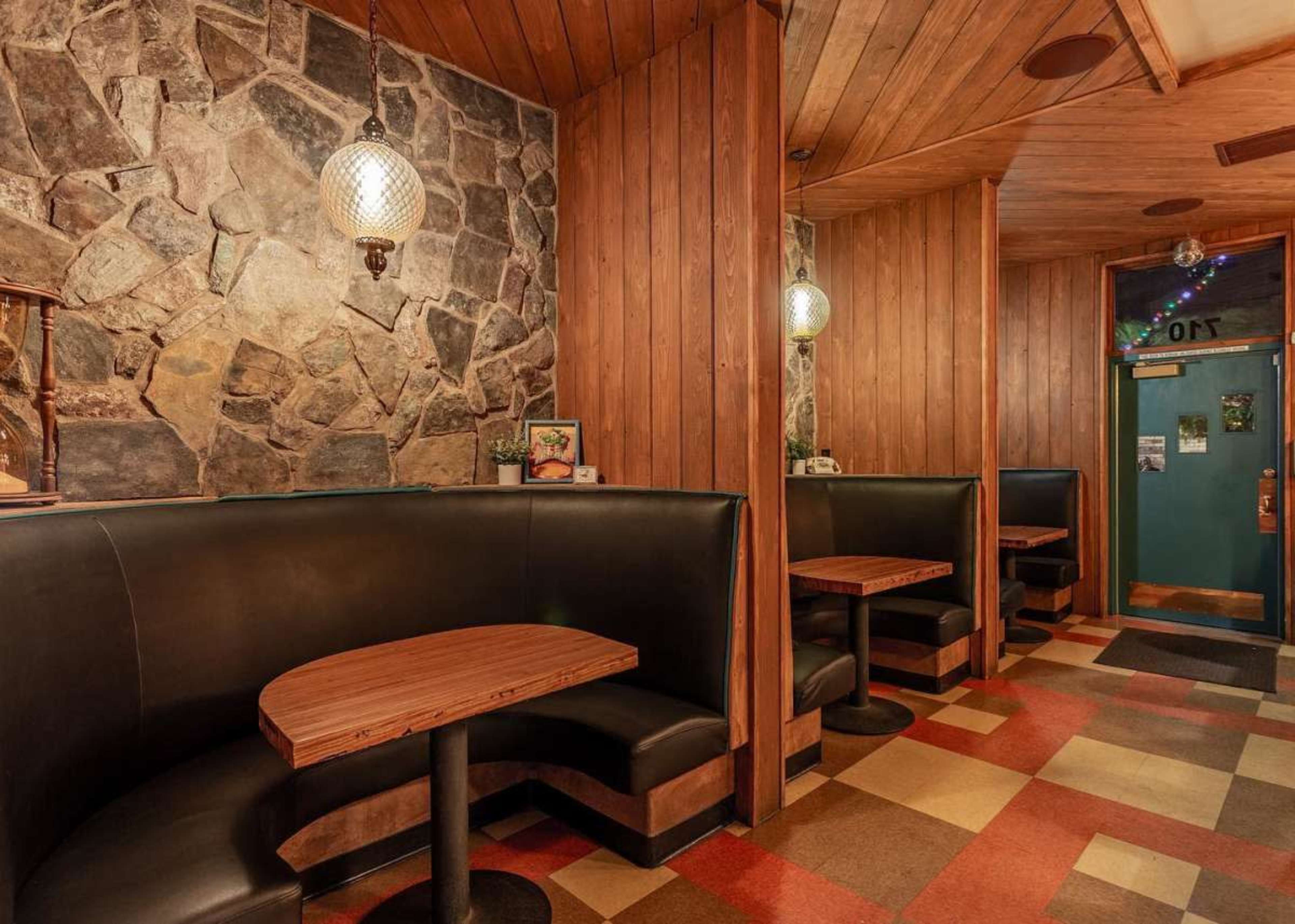 The image depicts a small restaurant interior with wooden walls, stone accents, and black booth seating arranged around wooden tables.