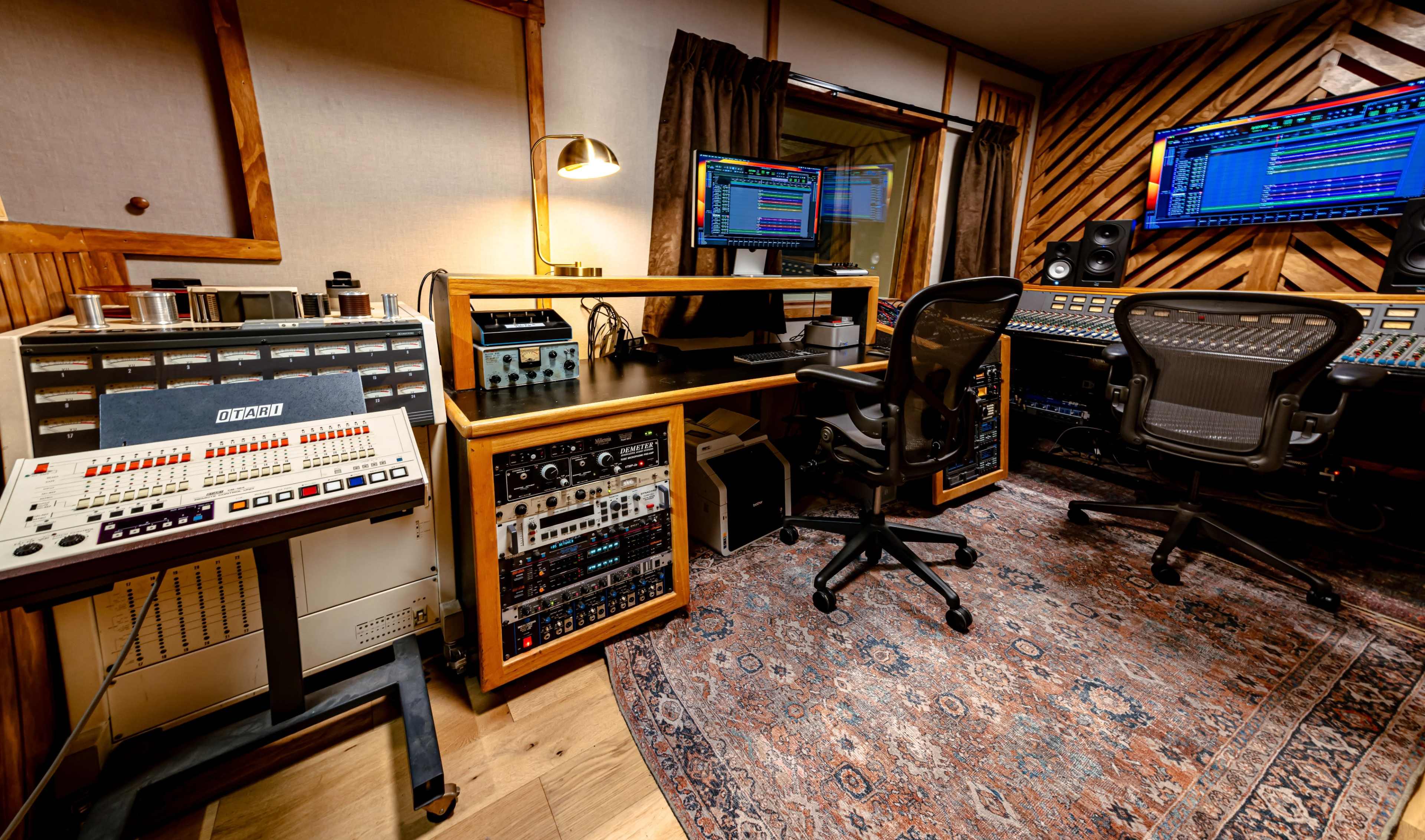 The image shows a music recording studio featuring recording equipment, a computer monitor, and mixing consoles arranged around a rug.