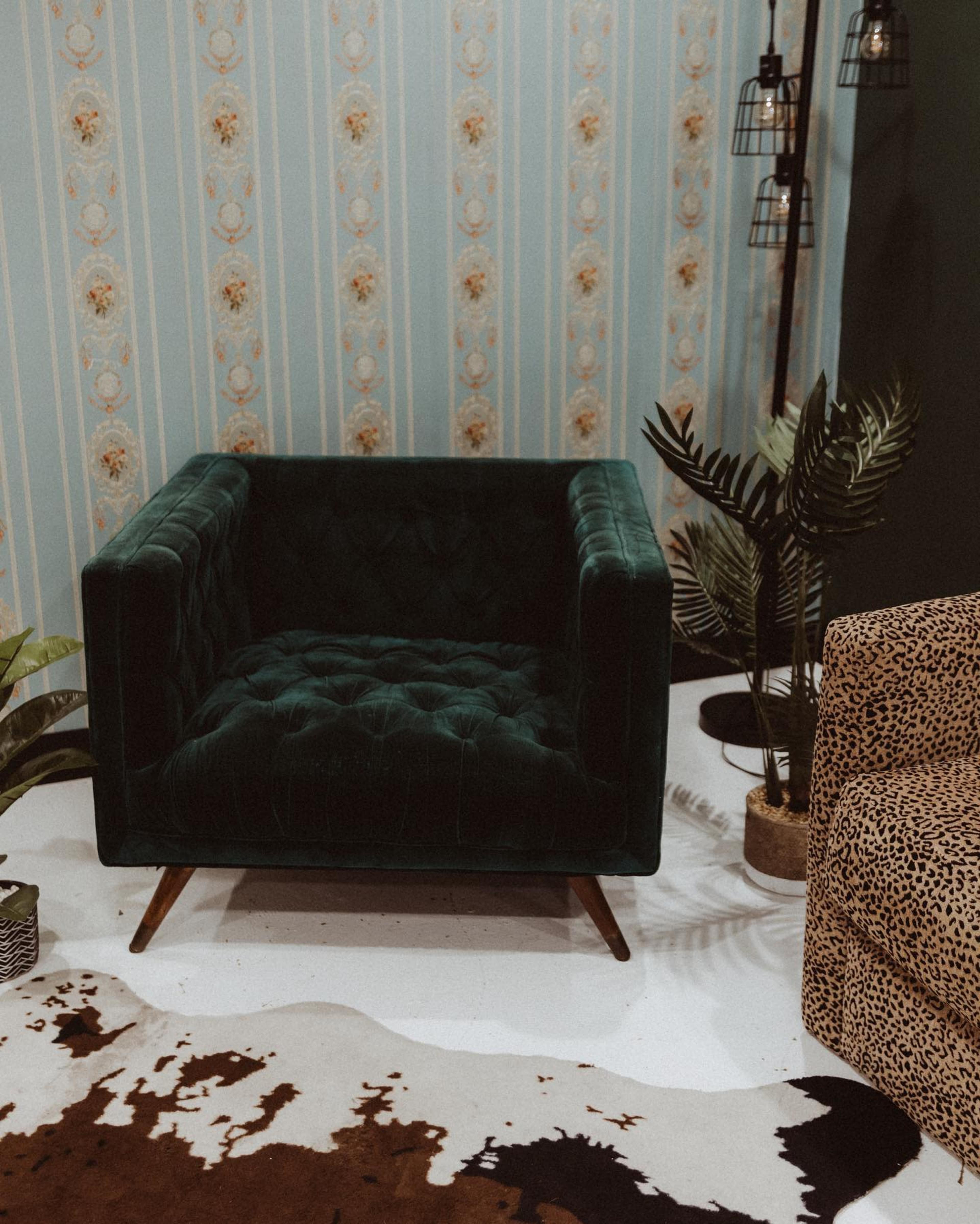 A green velvet armchair sits in a room with striped wallpaper and a patterned rug, surrounded by plants and a leopard print chair.