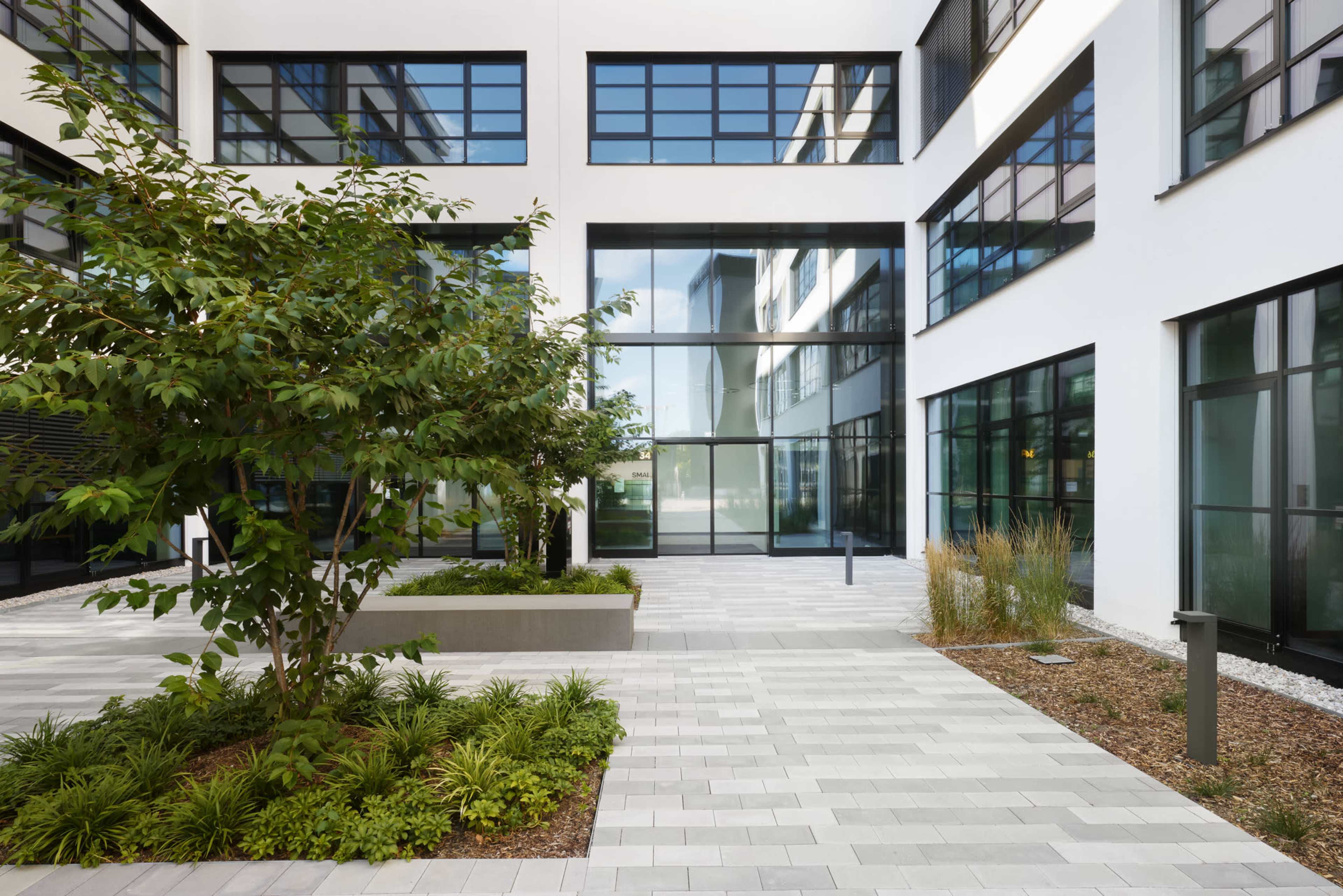 A modern courtyard features a central tree surrounded by tidy paving, glass walls, and landscaped areas.