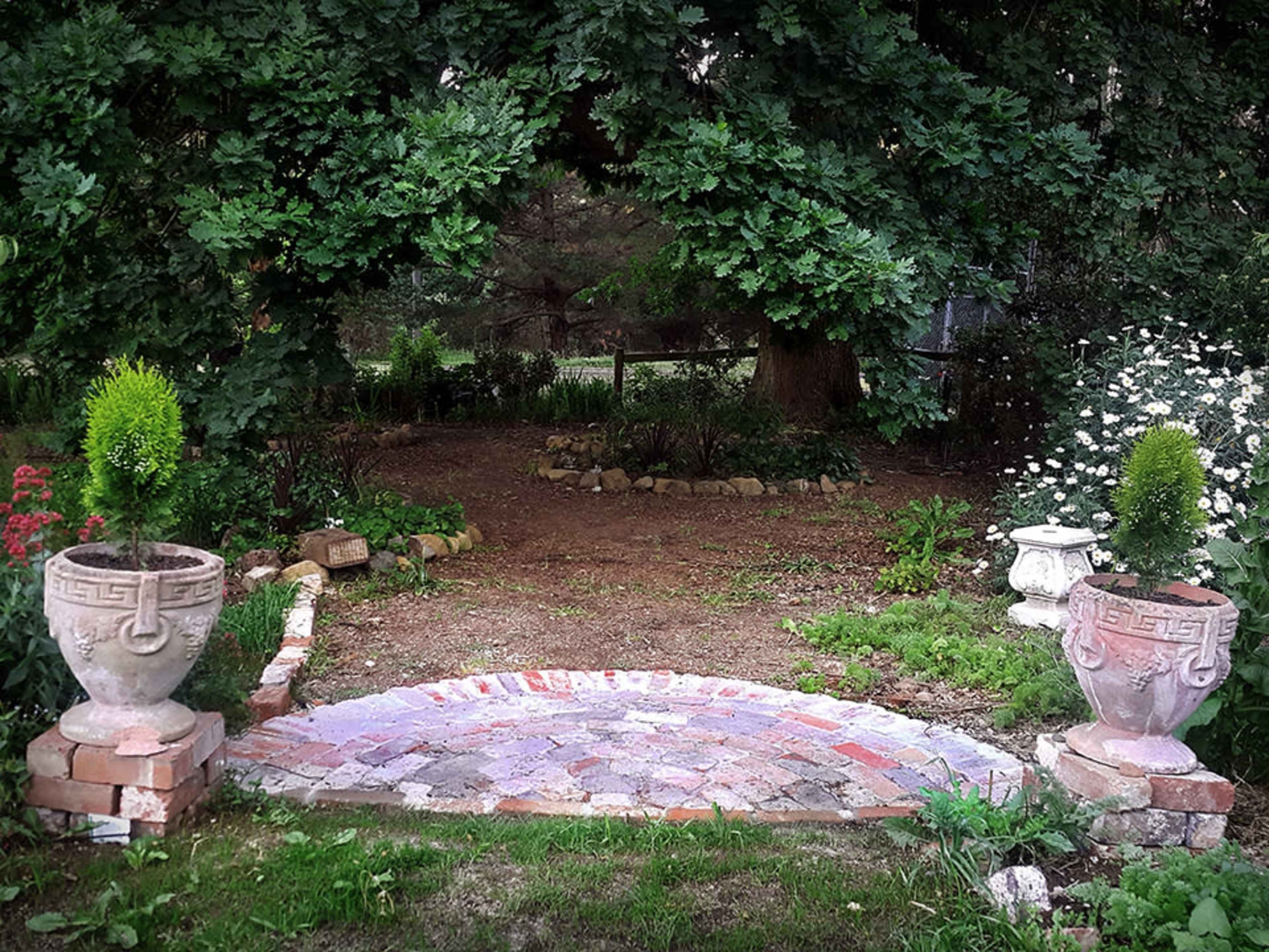 A stone path leads to a garden adorned with decorative urns and surrounded by greenery and flowering plants.