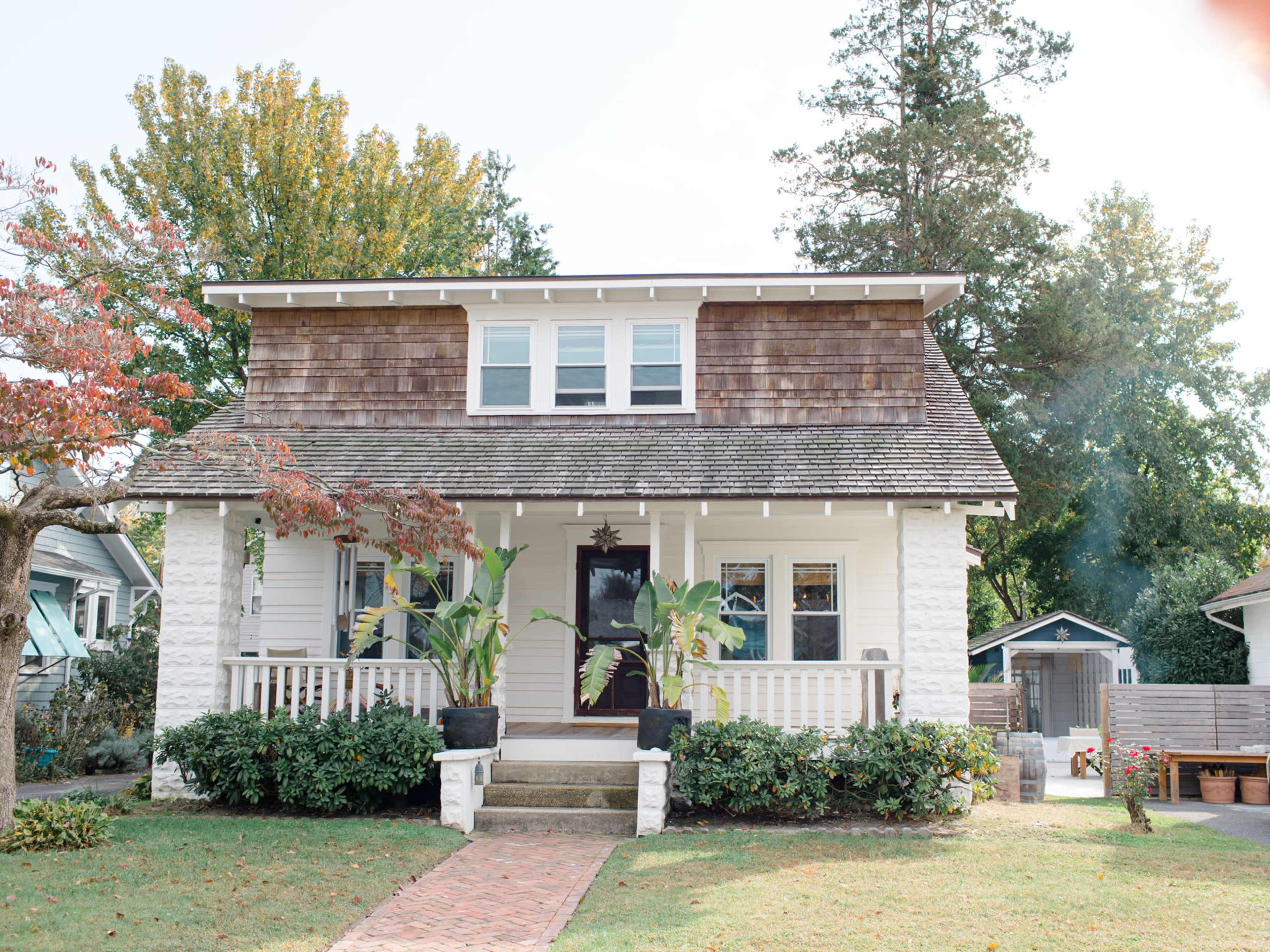 The image shows a two-story house with a shingle roof, a front porch, and two potted plants by the entrance.