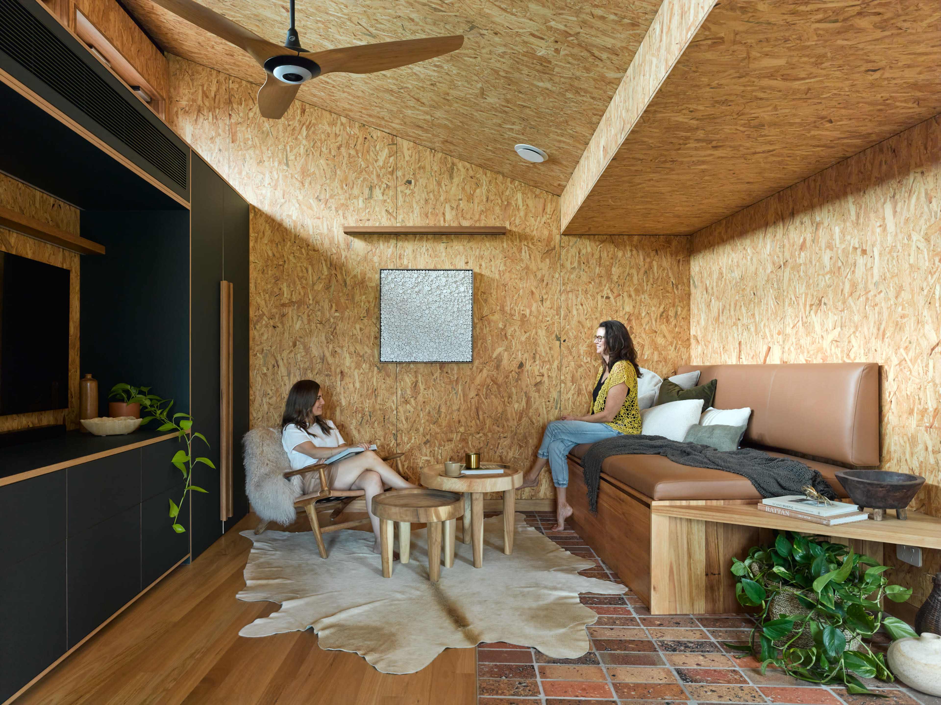 Two women are seated in a modern, wooden-paneled living room with a cowhide rug and plants, engaging in conversation.