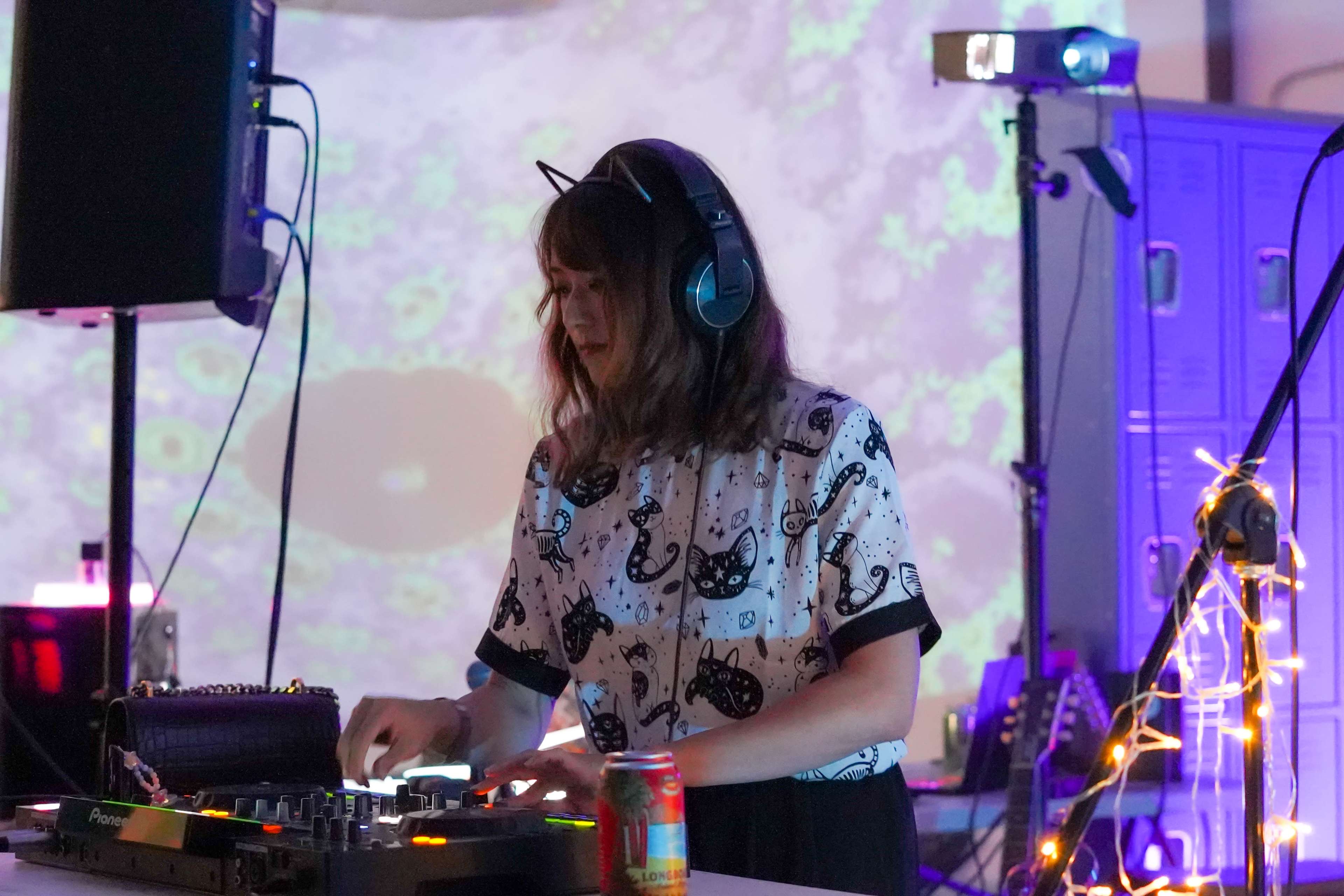 A DJ with cat-themed clothing is mixing music at a colorful event, with projected visuals and various equipment around her.