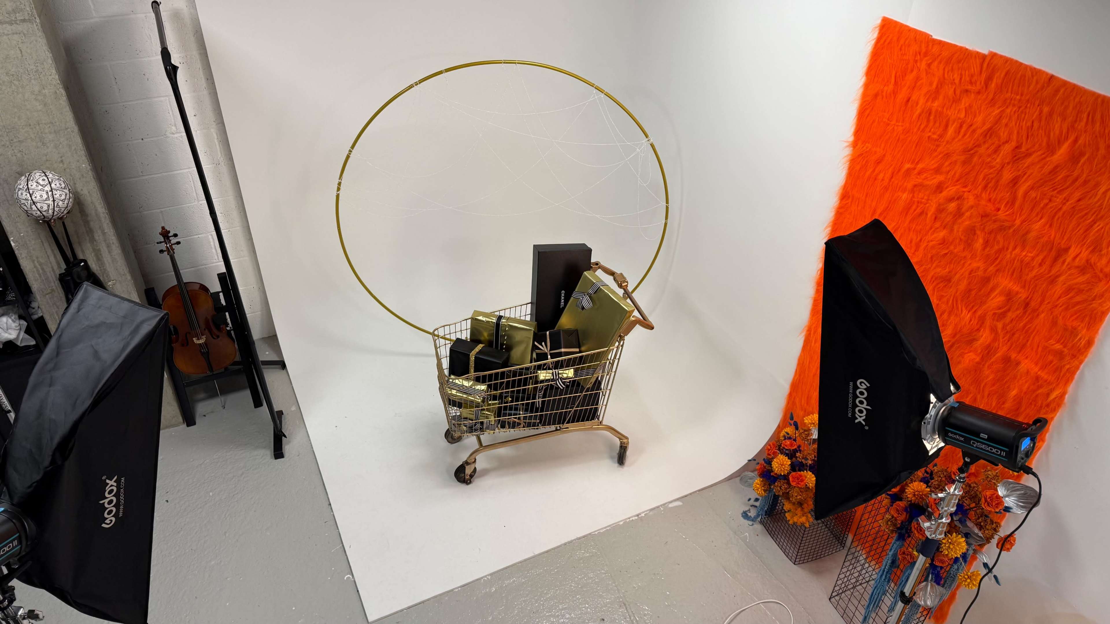 The image depicts a gold shopping cart filled with various items, positioned in a brightly lit studio with a vibrant orange backdrop and photography equipment nearby.