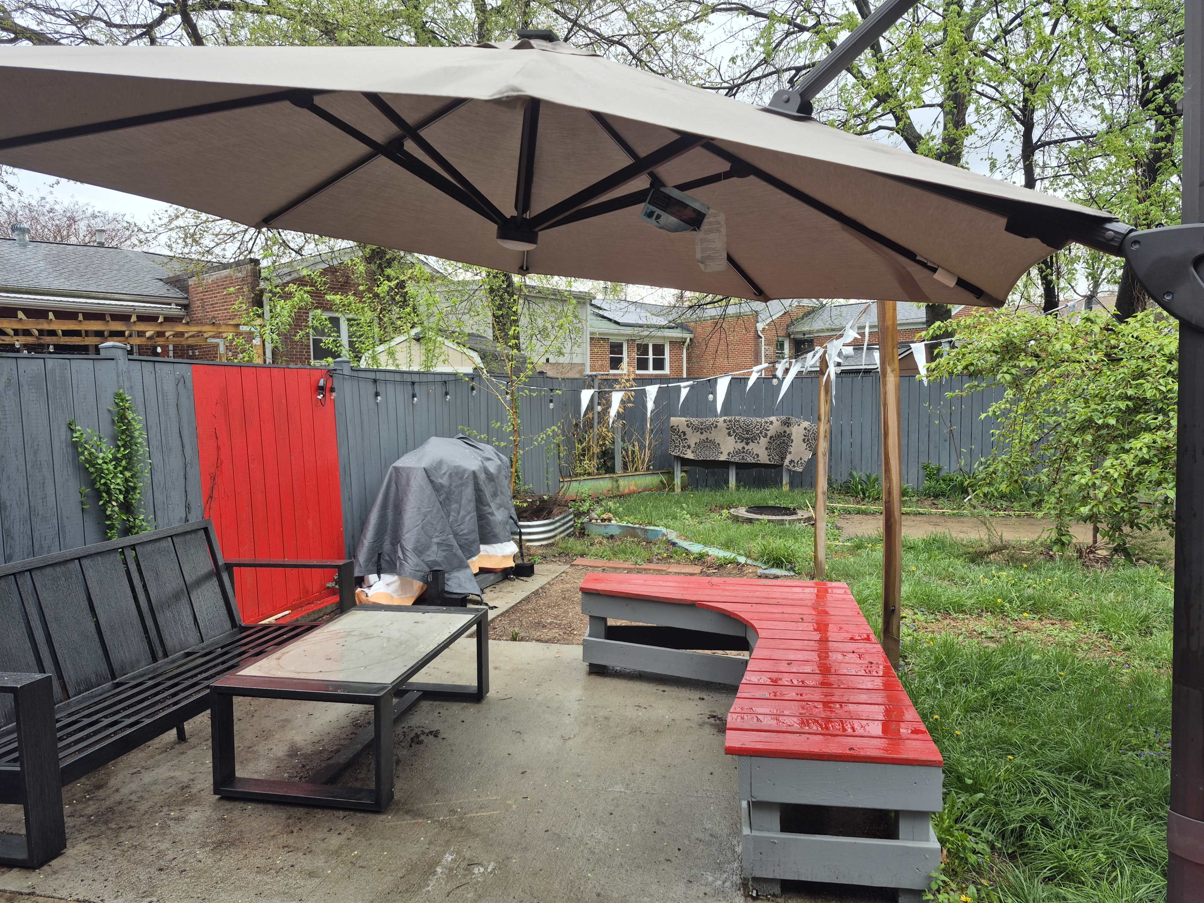The image shows a covered outdoor seating area with a red and black wooden bench, a glass table, and a nearby grill, surrounded by grass and fencing.