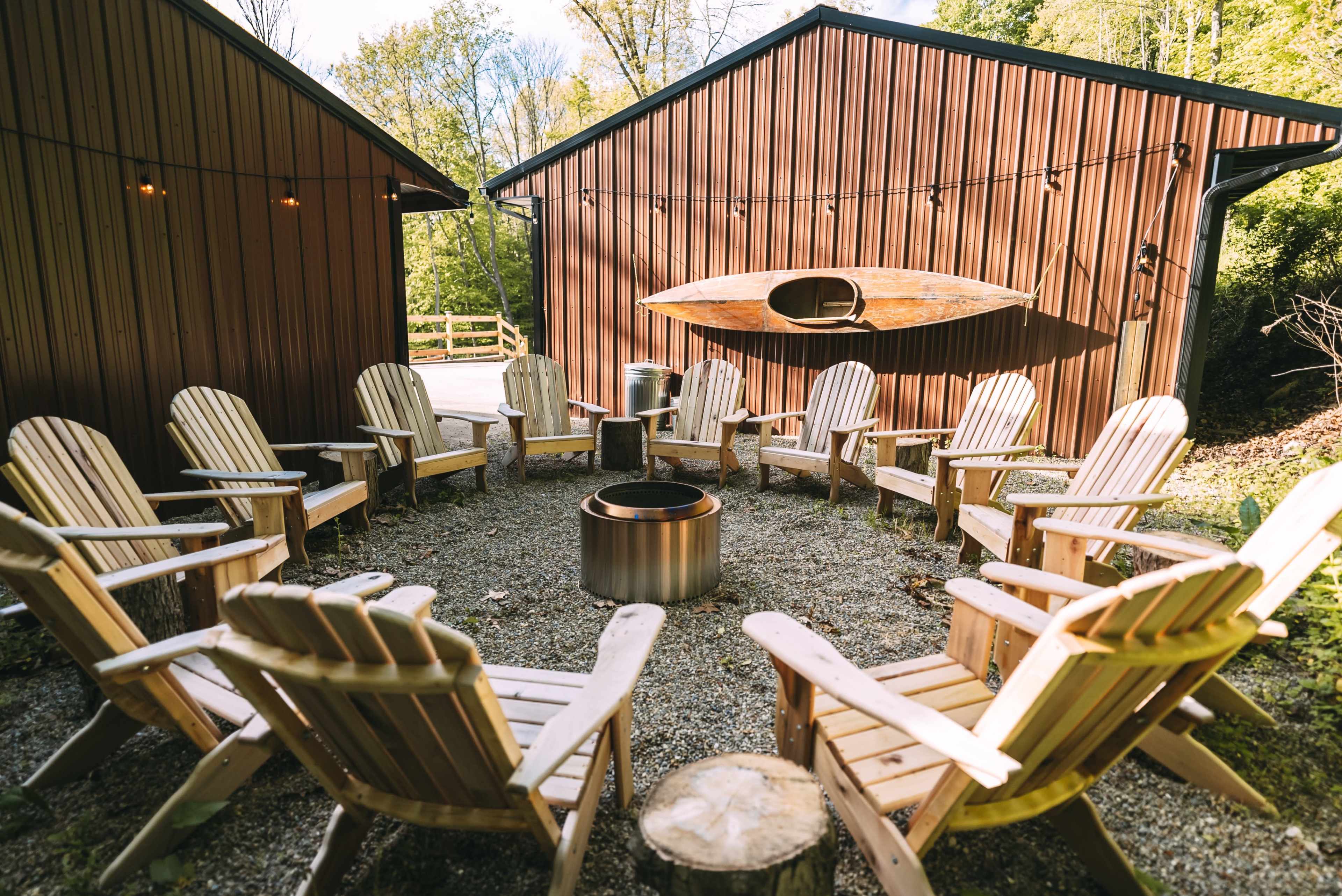 A circular arrangement of wooden chairs surrounds a fire pit between two brown buildings.