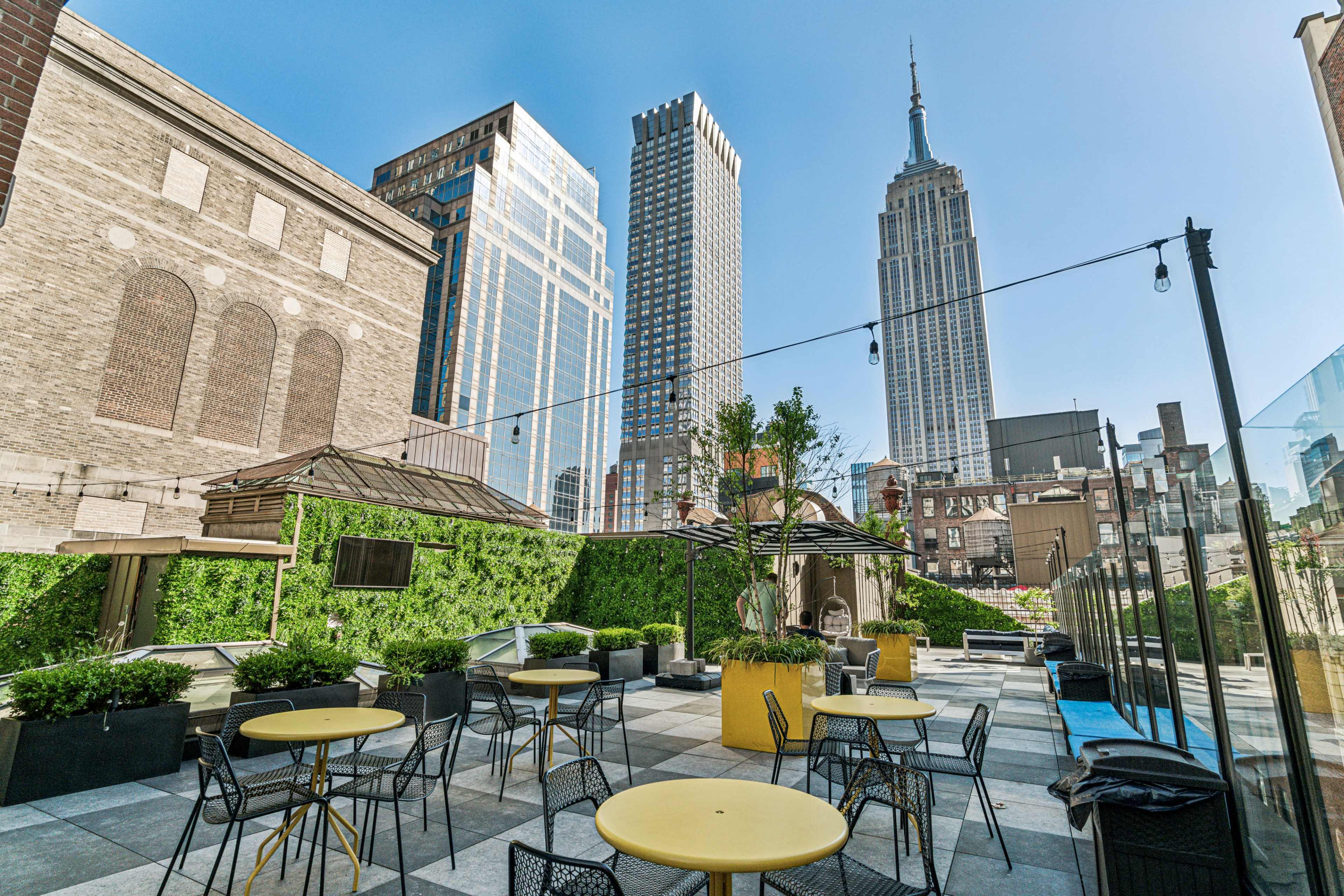 The image shows a rooftop terrace with yellow tables and chairs, surrounded by greenery, with the Empire State Building visible in the background.