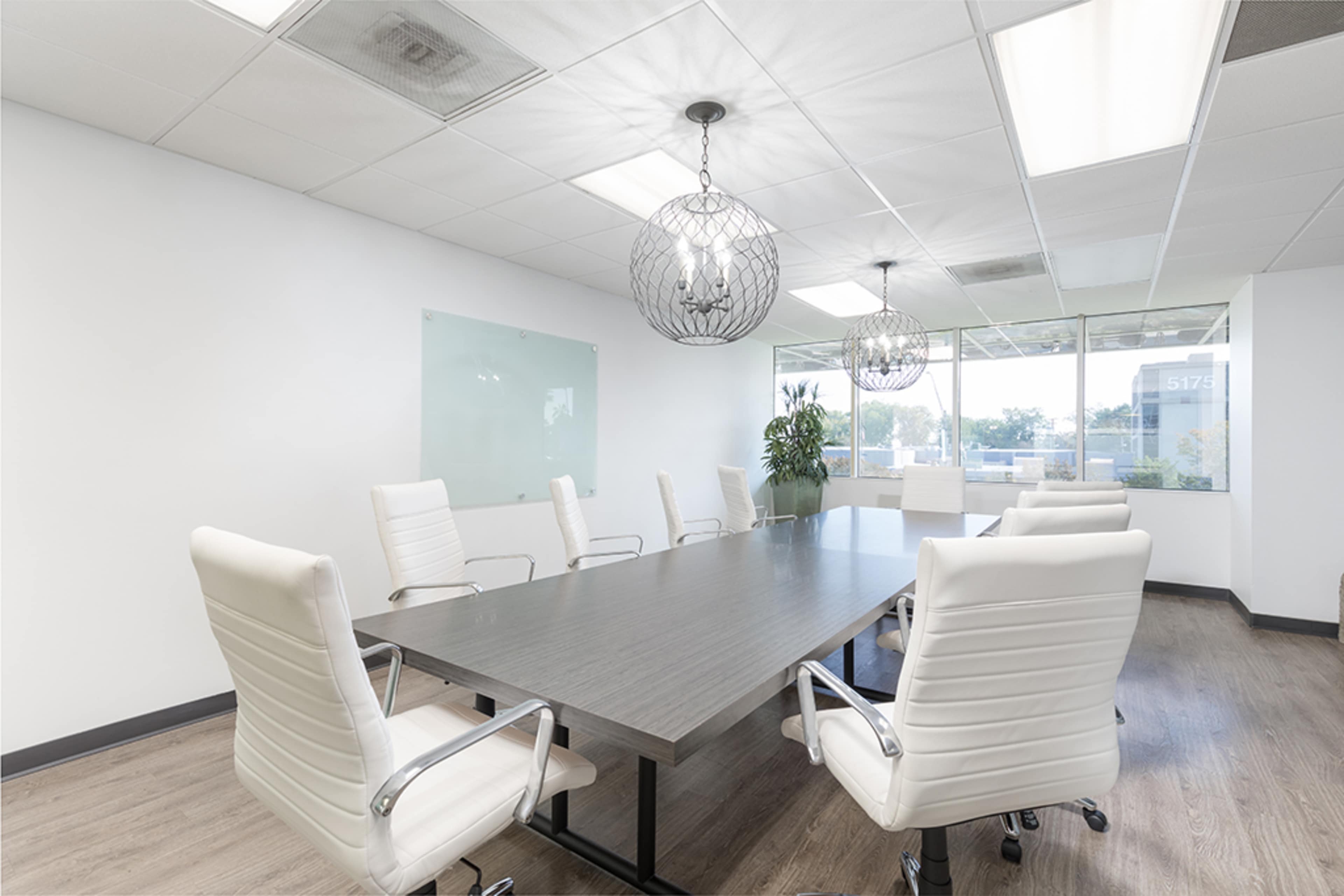 A modern conference room features a large wooden table surrounded by six white executive chairs, with pendant lighting and a window offering a view outside.
