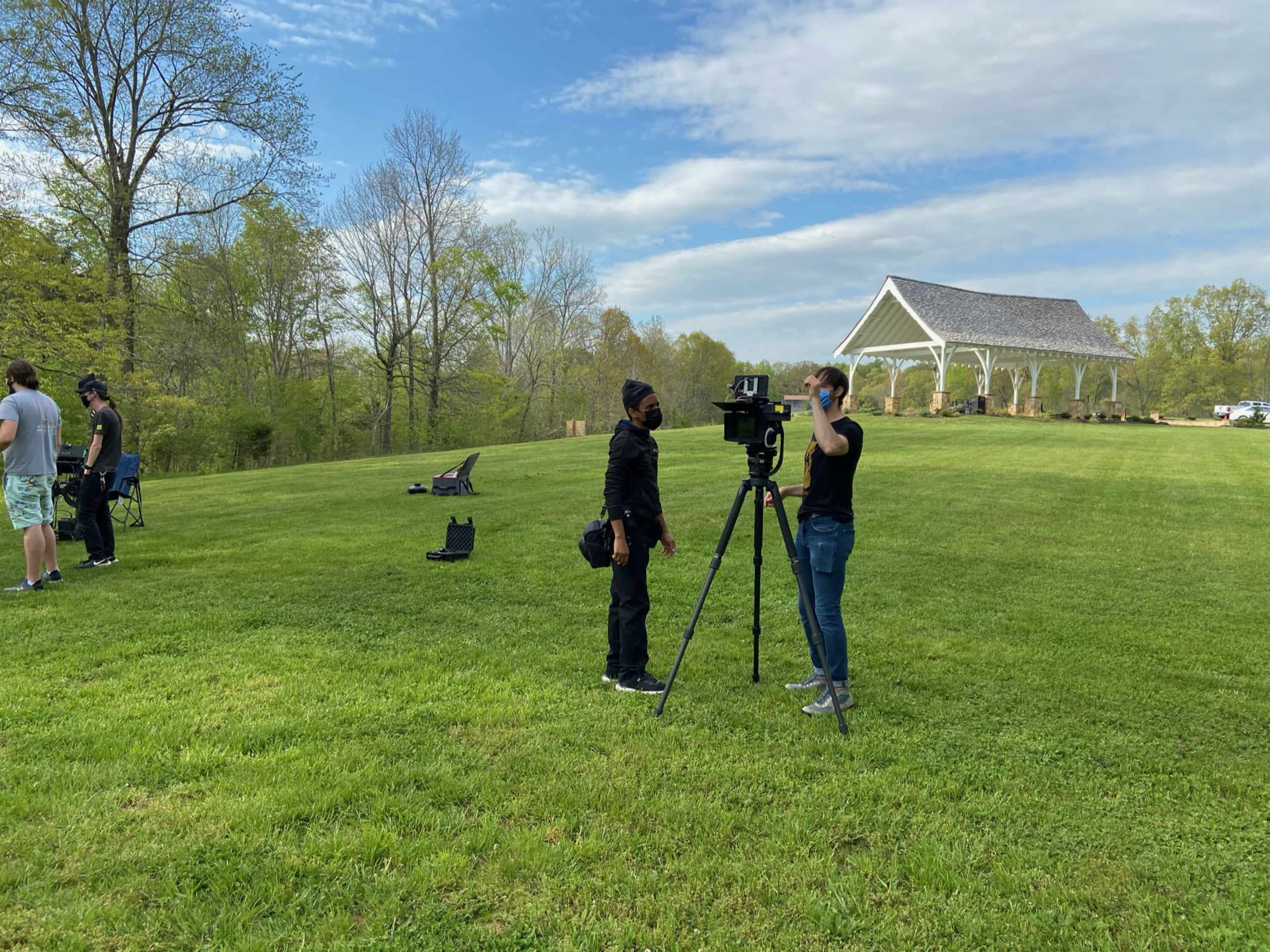 Two individuals set up equipment on a grassy field with a wooden pavilion in the background.