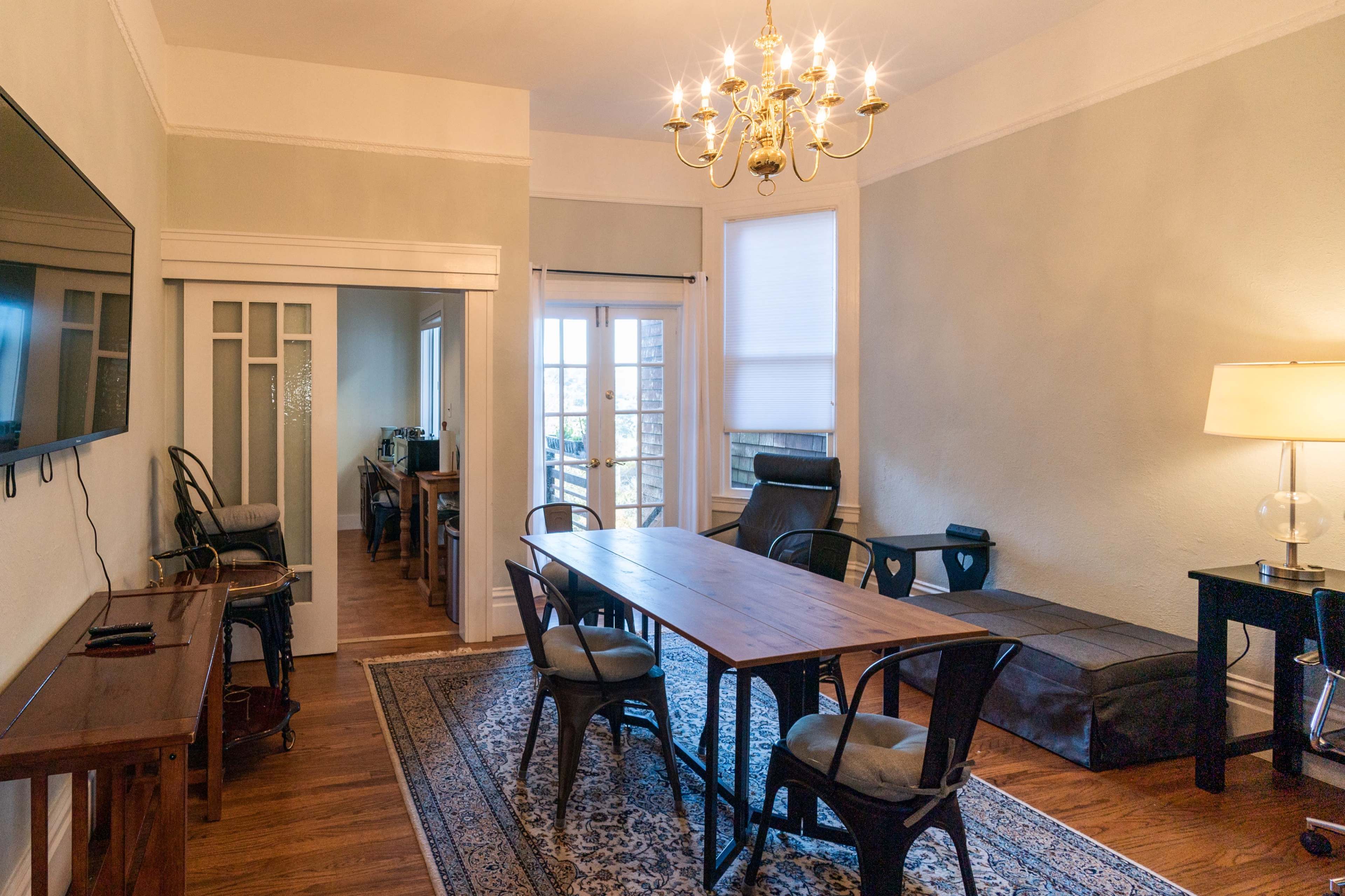 The image shows a furnished dining area with a wooden table surrounded by chairs, a television on the wall, and a chandelier overhead.