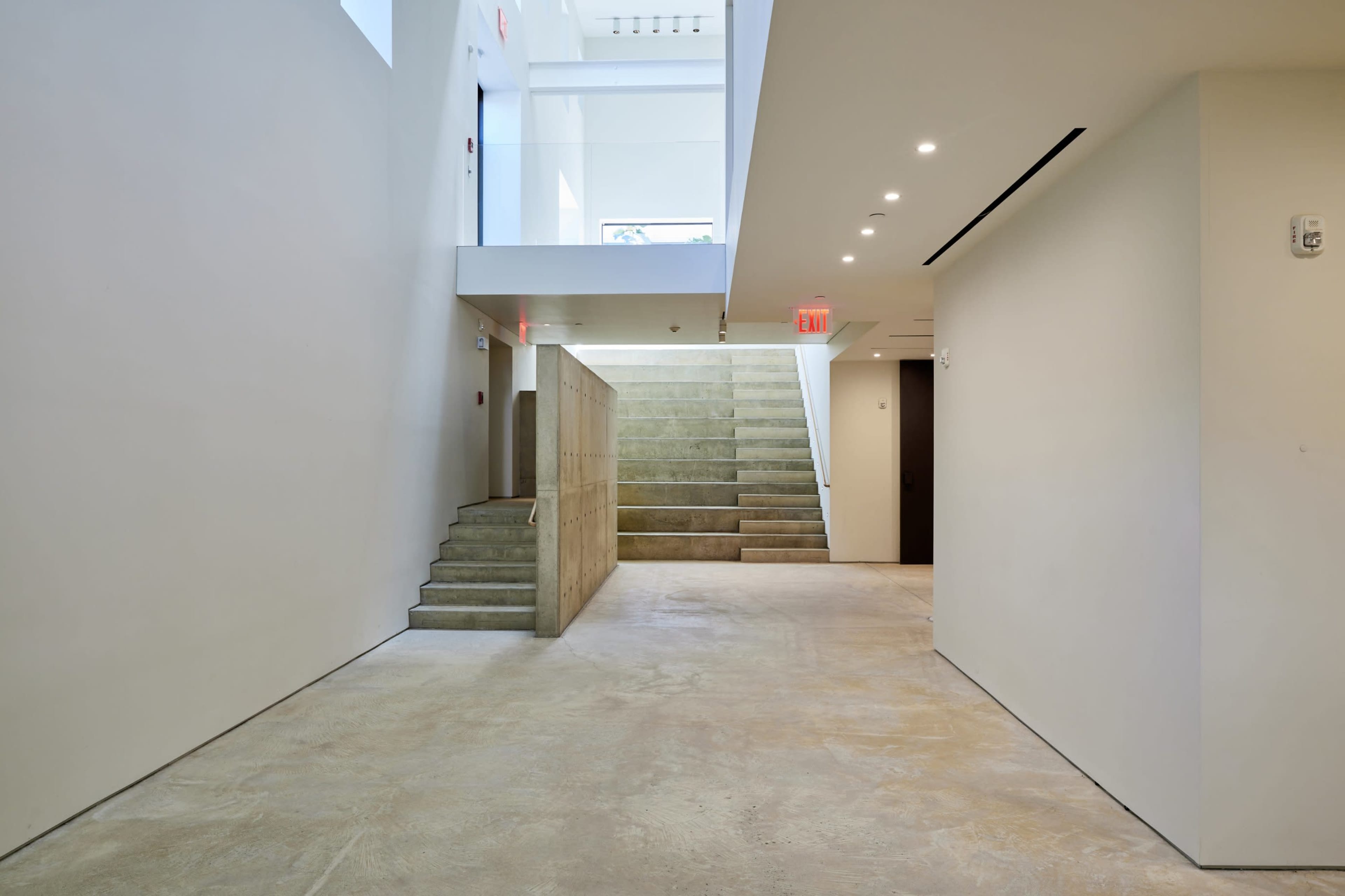 The image shows a modern interior space featuring a staircase on the left leading to an upper level, with minimalistic design and concrete flooring.