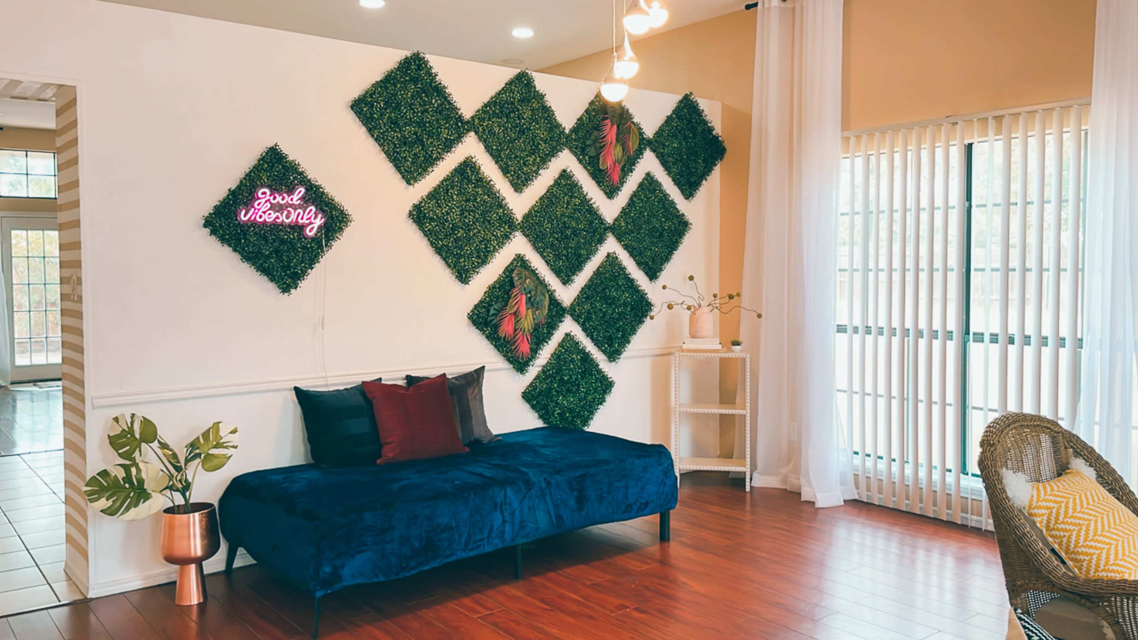 The image shows a living space featuring a blue couch, decorative wall greenery arranged in a diamond pattern, and a neon sign that reads "Good Vibes Only."