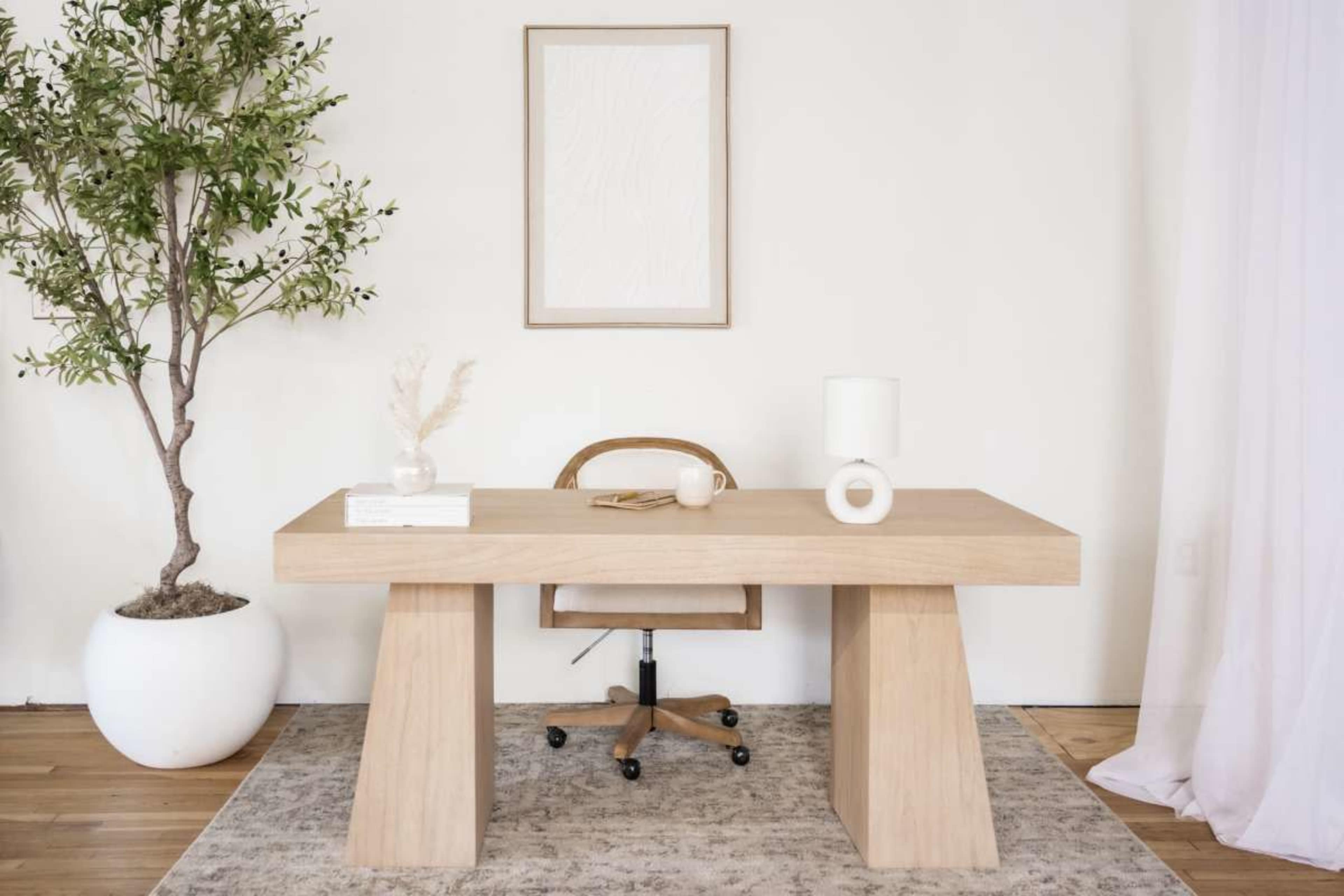 A modern workspace features a wooden desk with a minimalist chair, a small plant, and decorative items on a textured rug.