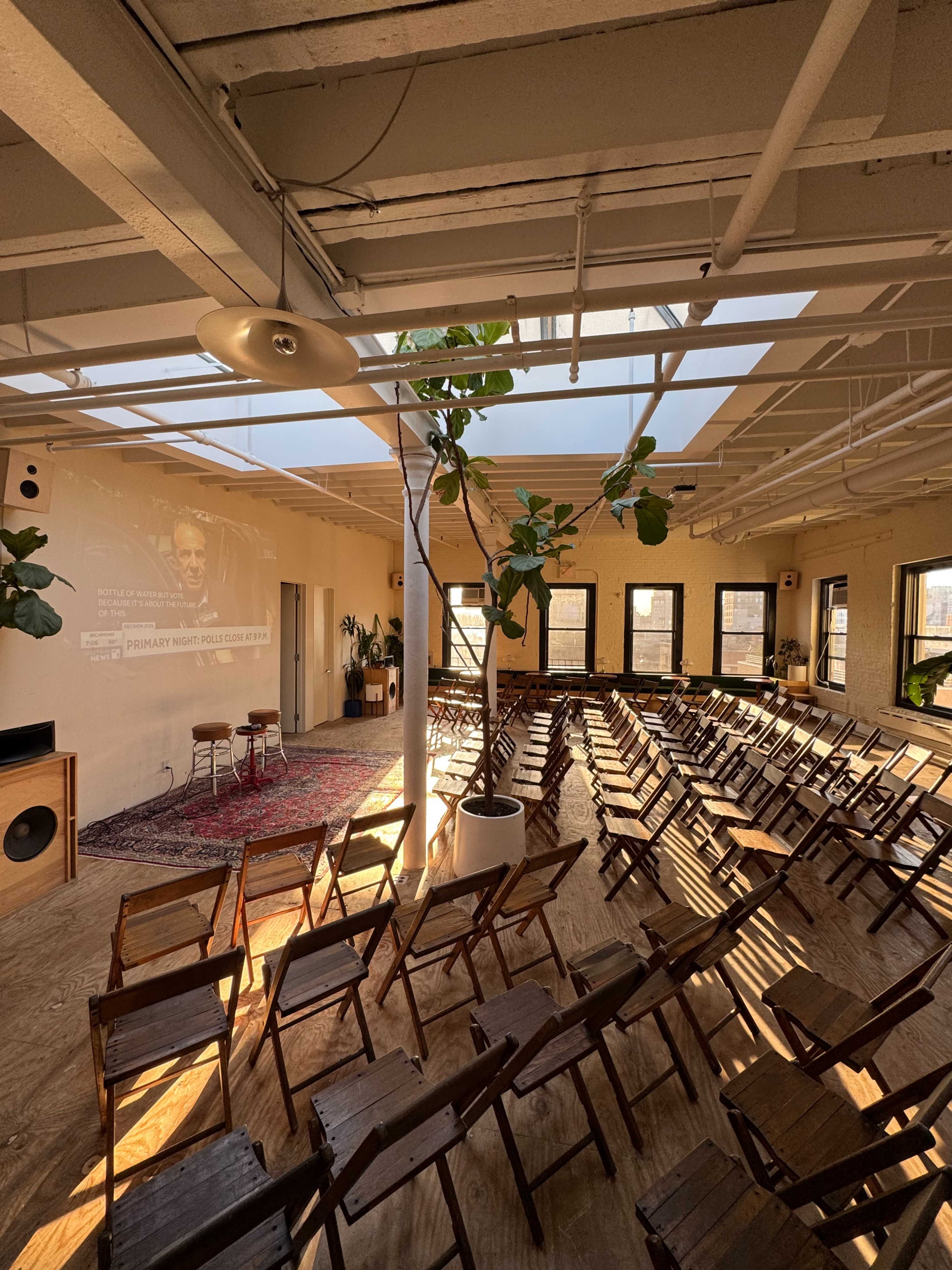 The image shows a spacious, well-lit room set up for an event, featuring rows of wooden chairs, a large screen displaying a presentation, and plants scattered throughout.