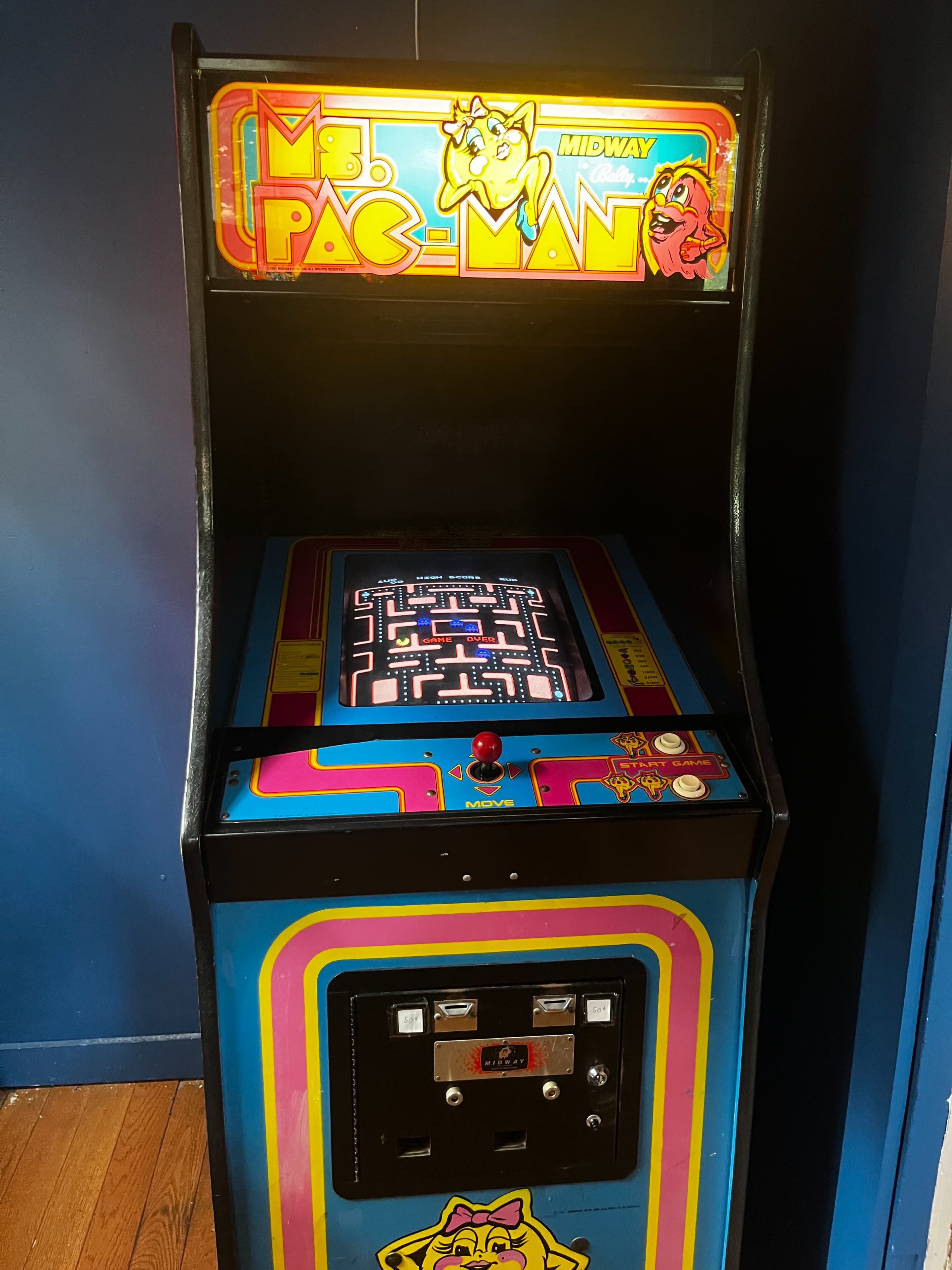 The image shows a classic Ms. Pac-Man arcade game cabinet positioned against a blue wall.