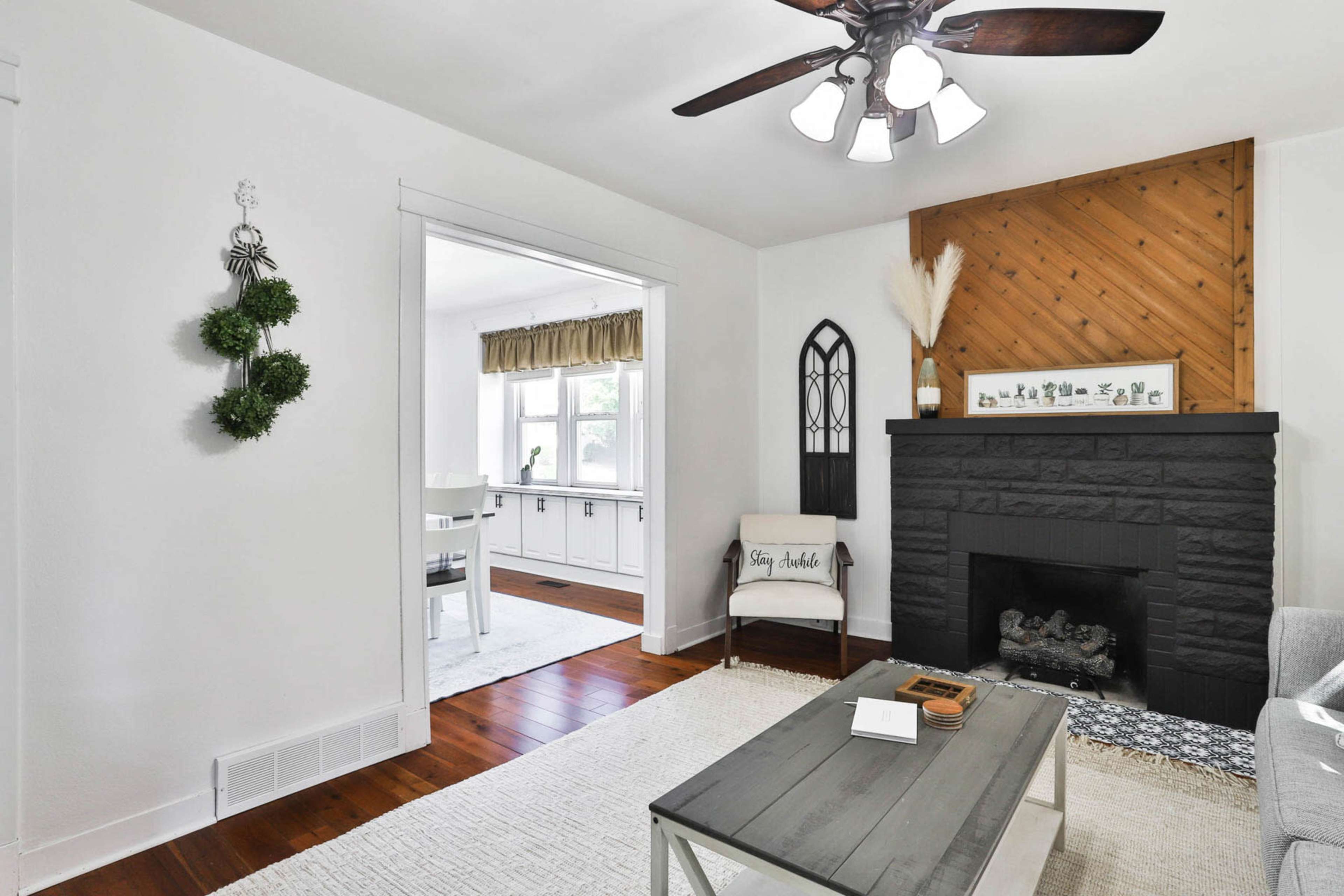 A cozy living room with a black stone fireplace, a wooden ceiling fan, and a view into a bright, adjacent room with white cabinetry.