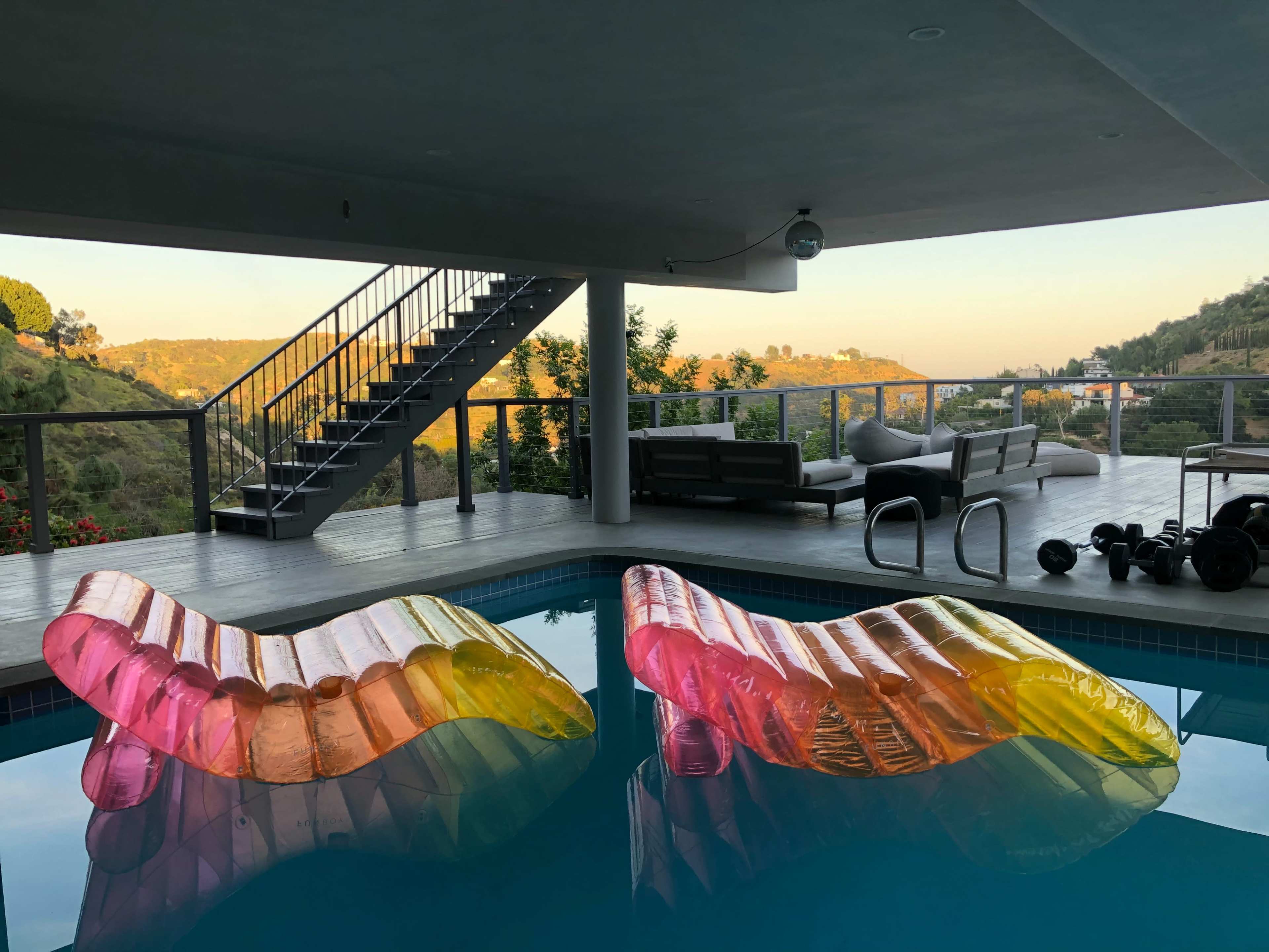 Two colorful inflatable loungers in a swimming pool, with a modern patio area and a staircase in the background overlooking a hillside landscape.