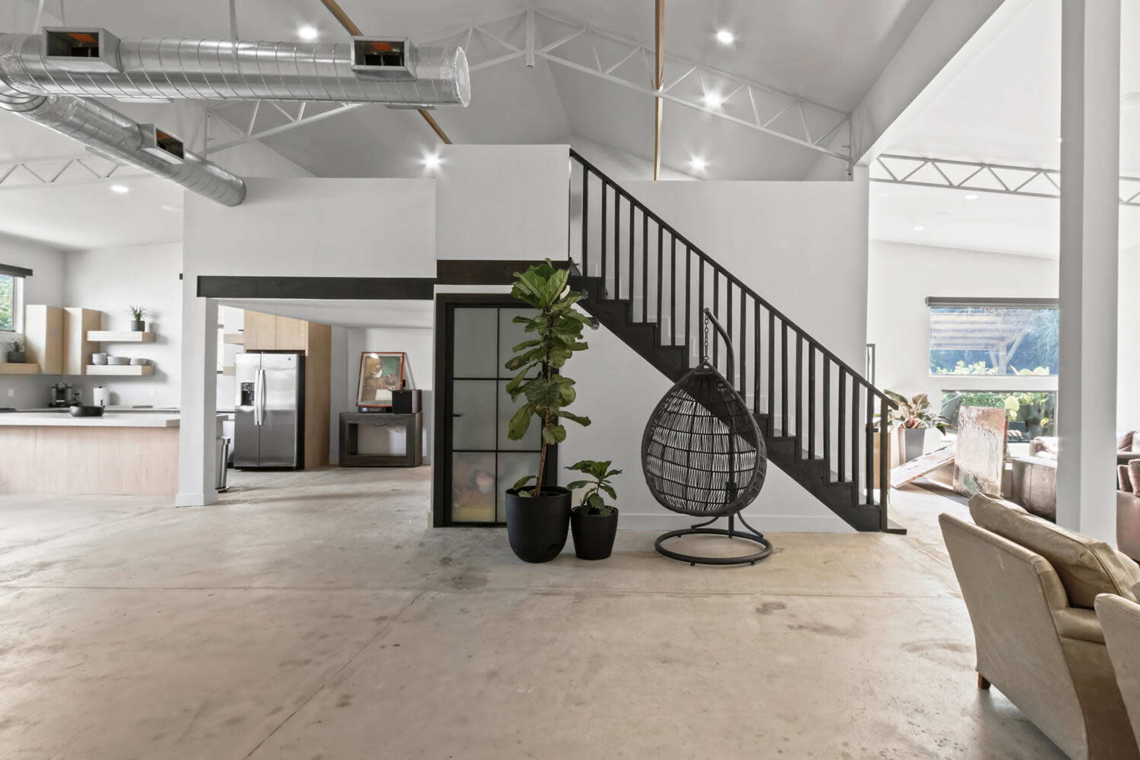 The image shows an open-concept living space featuring a staircase with a black railing, a hanging chair, and large windows that provide natural light.