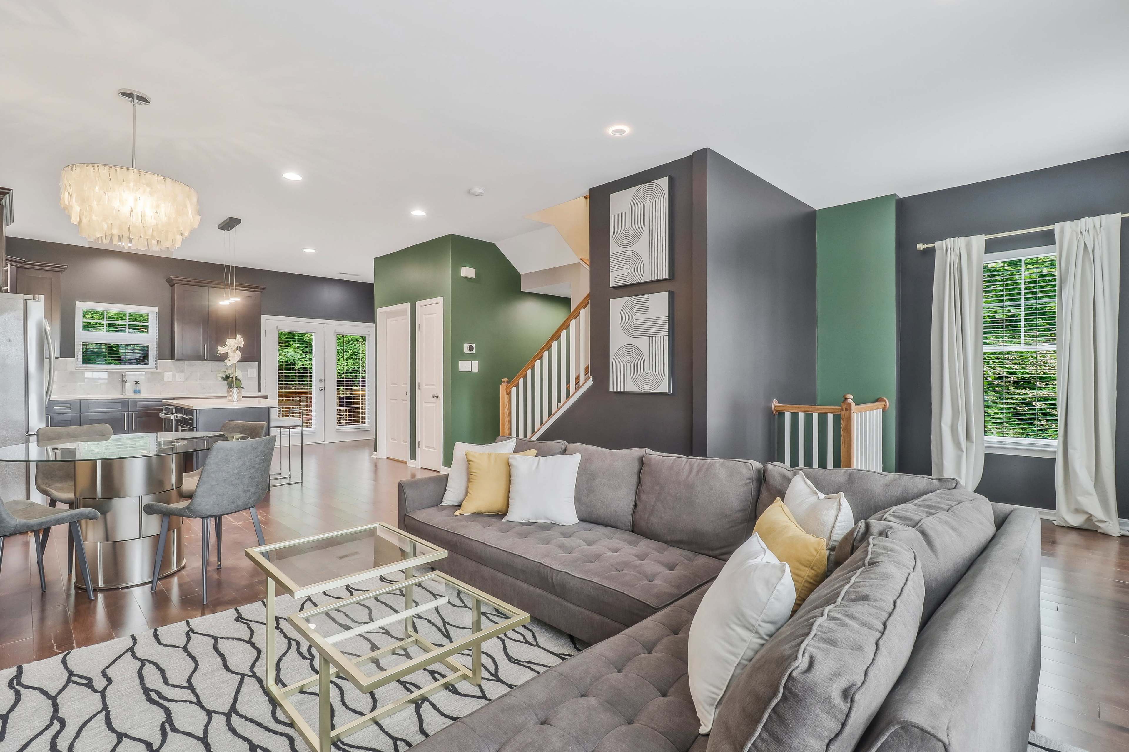 A modern living room features a gray sectional sofa, a glass coffee table, and a dining area with a chandelier, set against dark green walls and large windows.