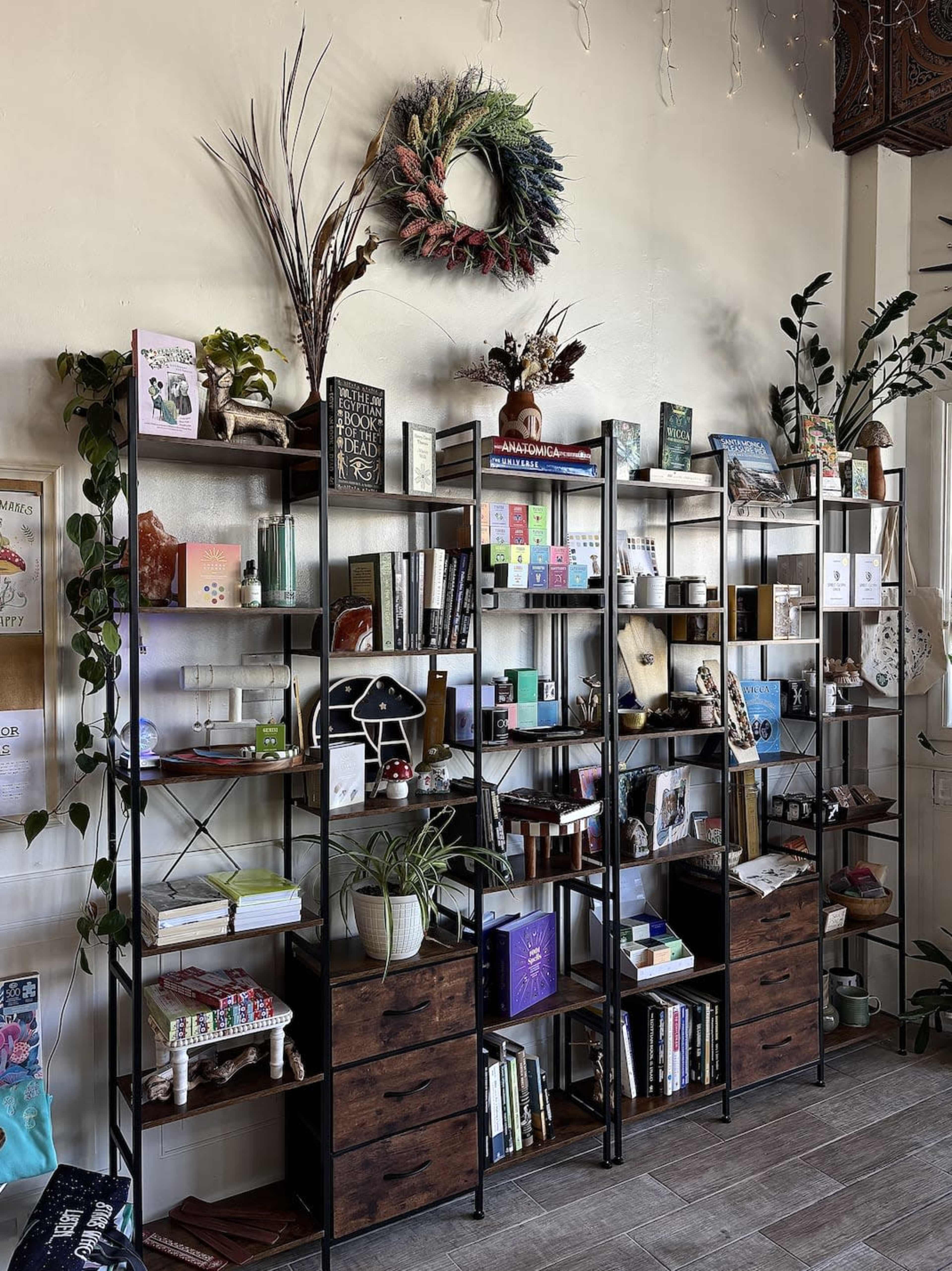 The image shows a wall-mounted display of open shelving filled with books, plants, and decorative items, along with a wreath hanging above.