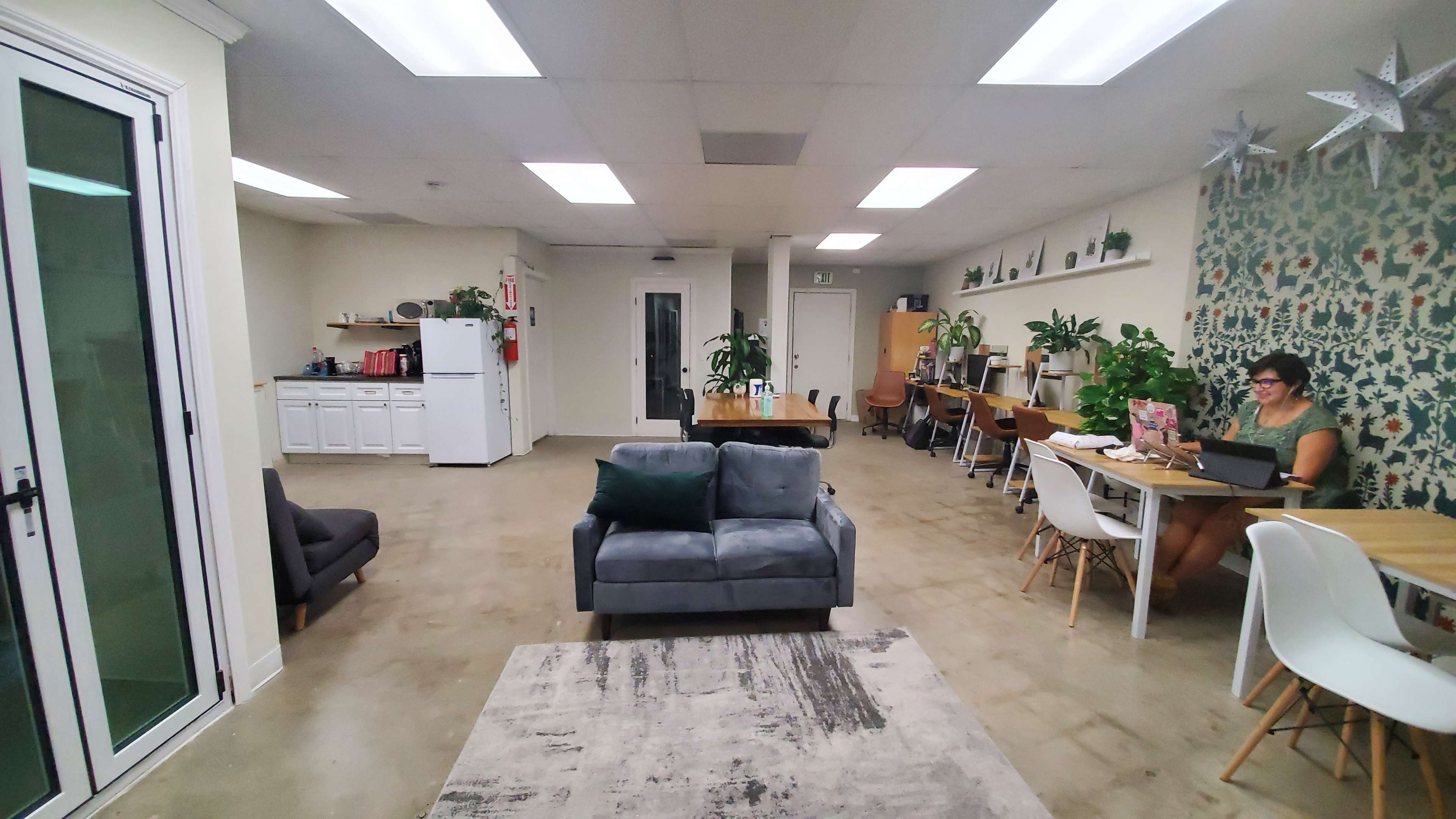 The image shows a modern office space featuring a kitchen area, a seating area with a gray couch, and several workstations with plants and a woman typing on a laptop.