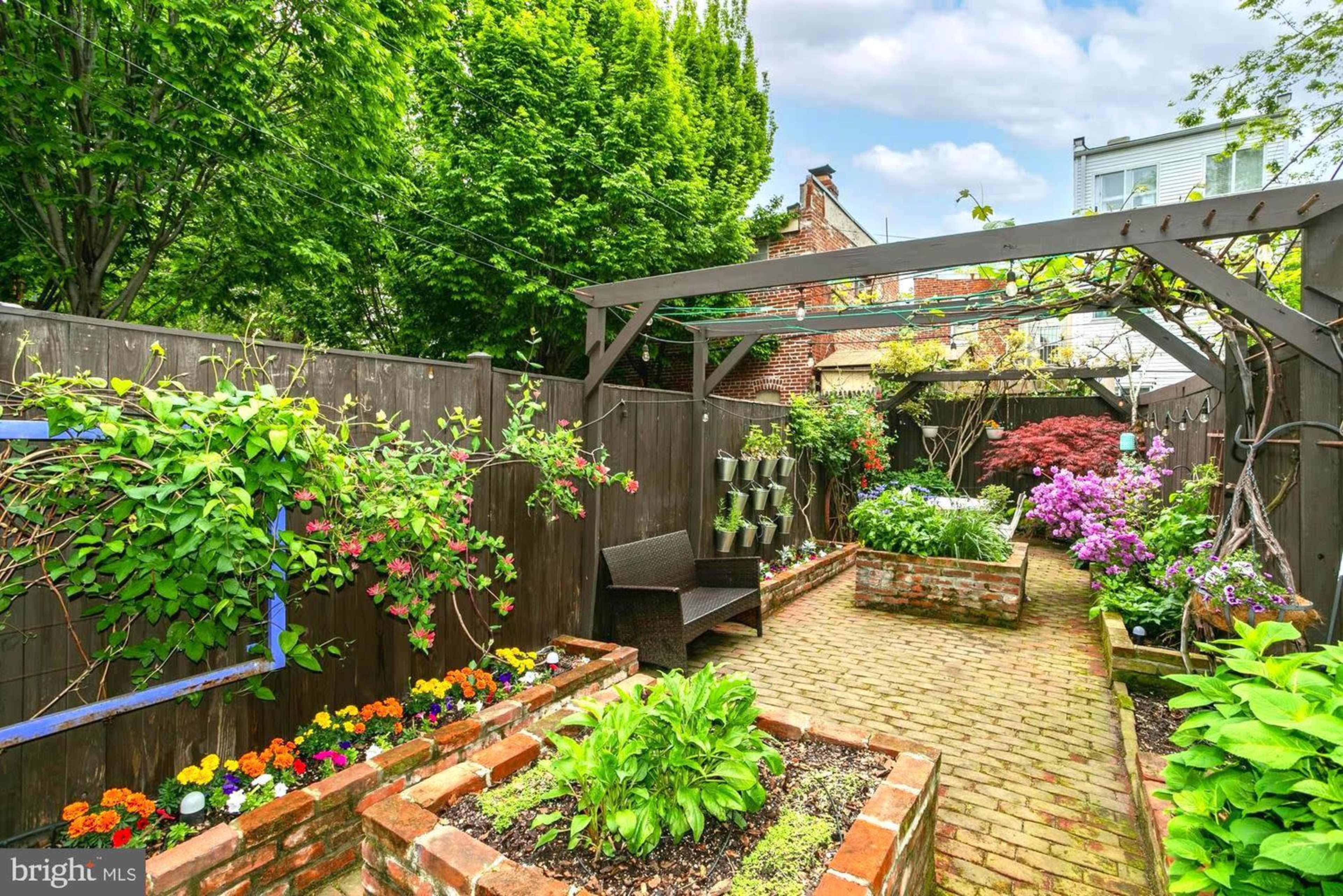 The image shows a neatly arranged garden path lined with colorful flowers and plants, bordered by wooden fences and trellises.