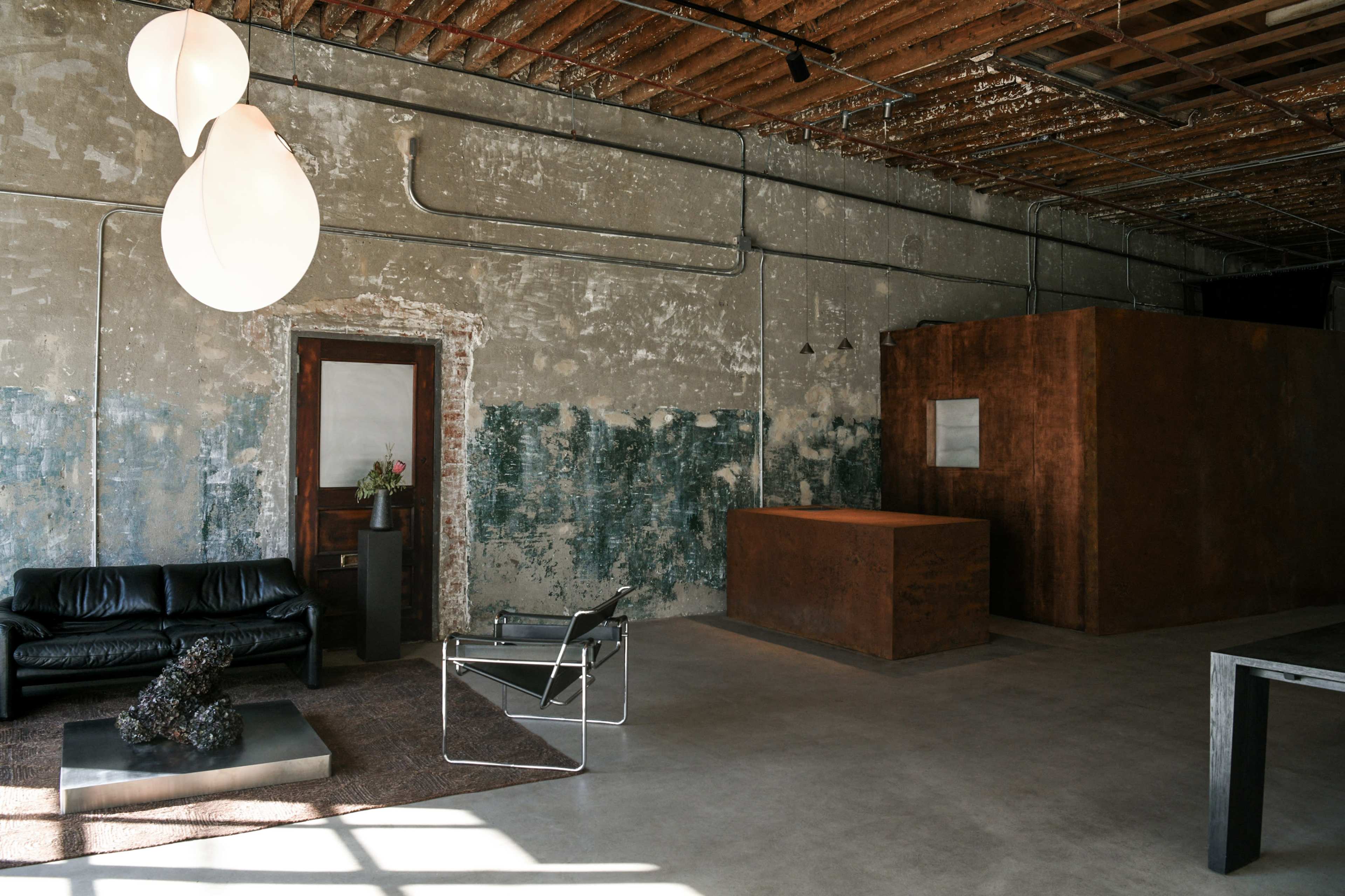 The image depicts an industrial-style interior featuring a black leather sofa, a modern chair, and a large copper-colored structure against a textured, peeling wall.