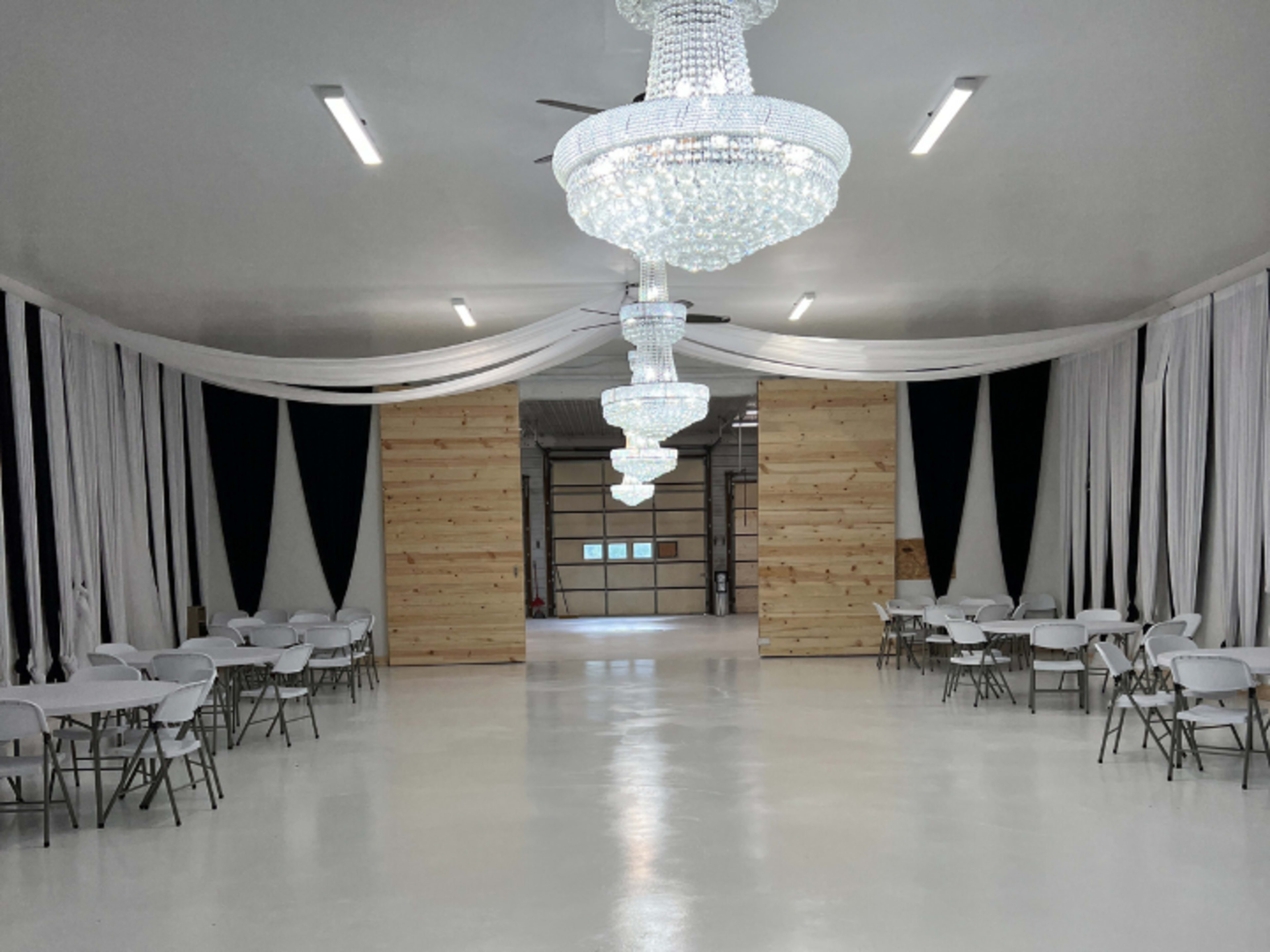 The image shows a spacious, well-lit interior with white walls, elegant chandeliers, and rows of folding chairs set up around white tables, framed by fabric drapes.