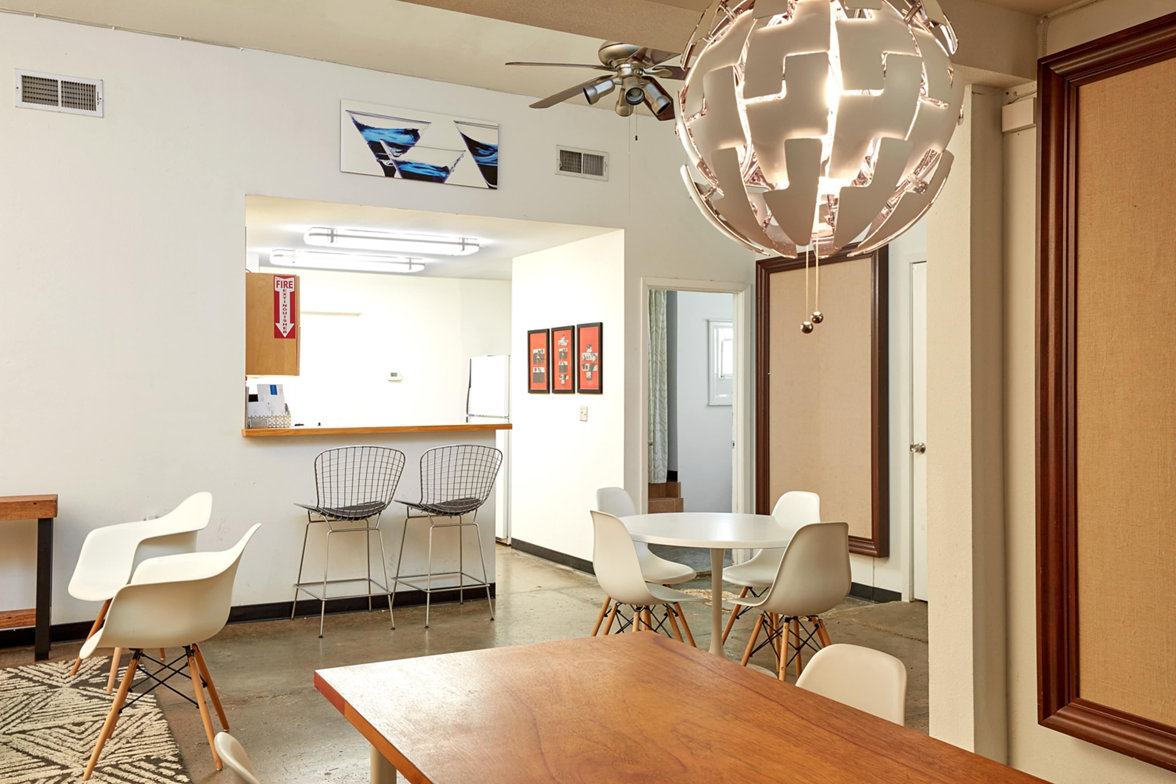 A modern, open-plan interior space featuring a dining area with a wooden table and white chairs, a round table in the foreground, and a kitchen area in the background with bar stools and framed artwork on the walls.