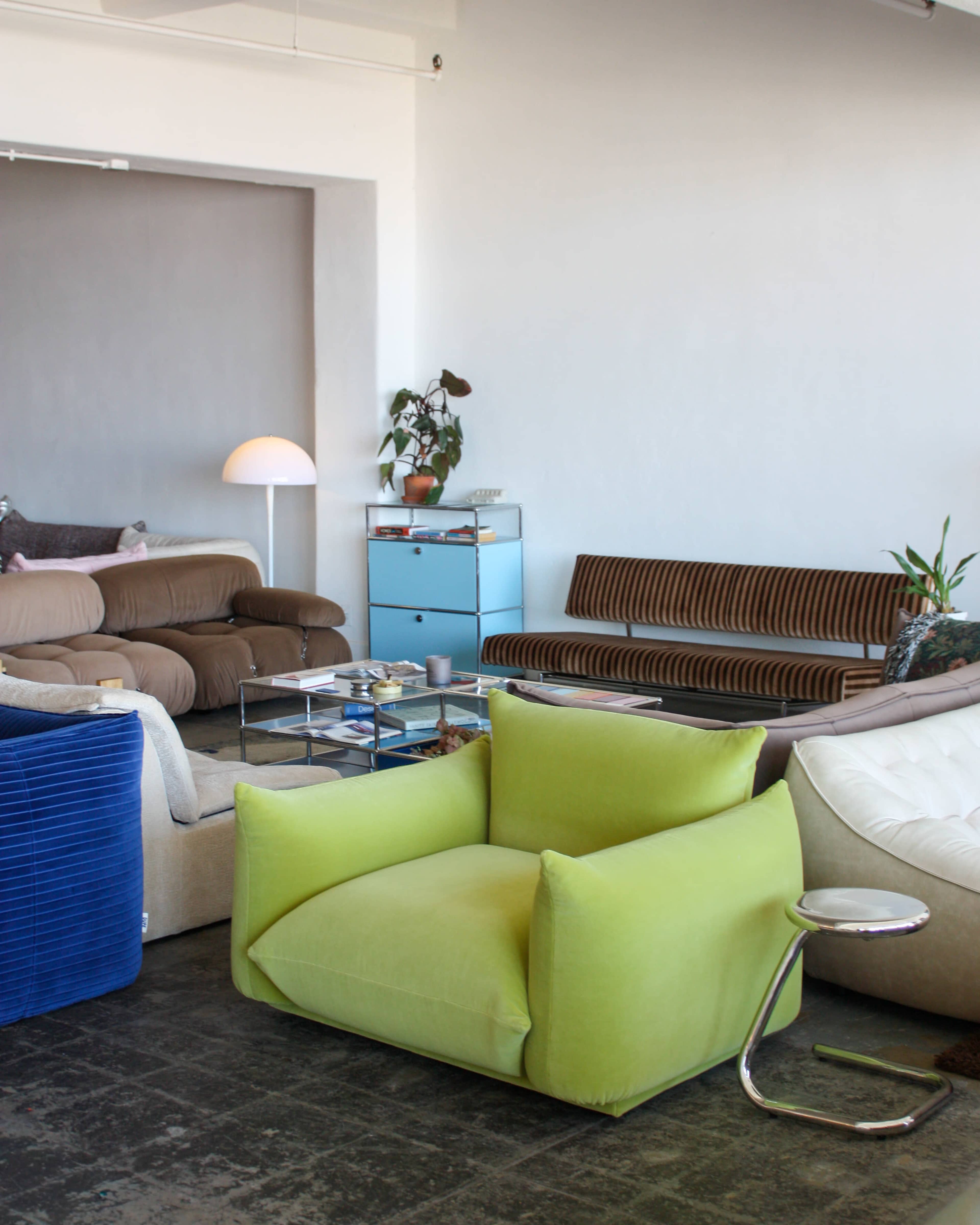The image shows a living room with a variety of seating options, including a green chair, several couches, a glass coffee table, and a lamp in the background.
