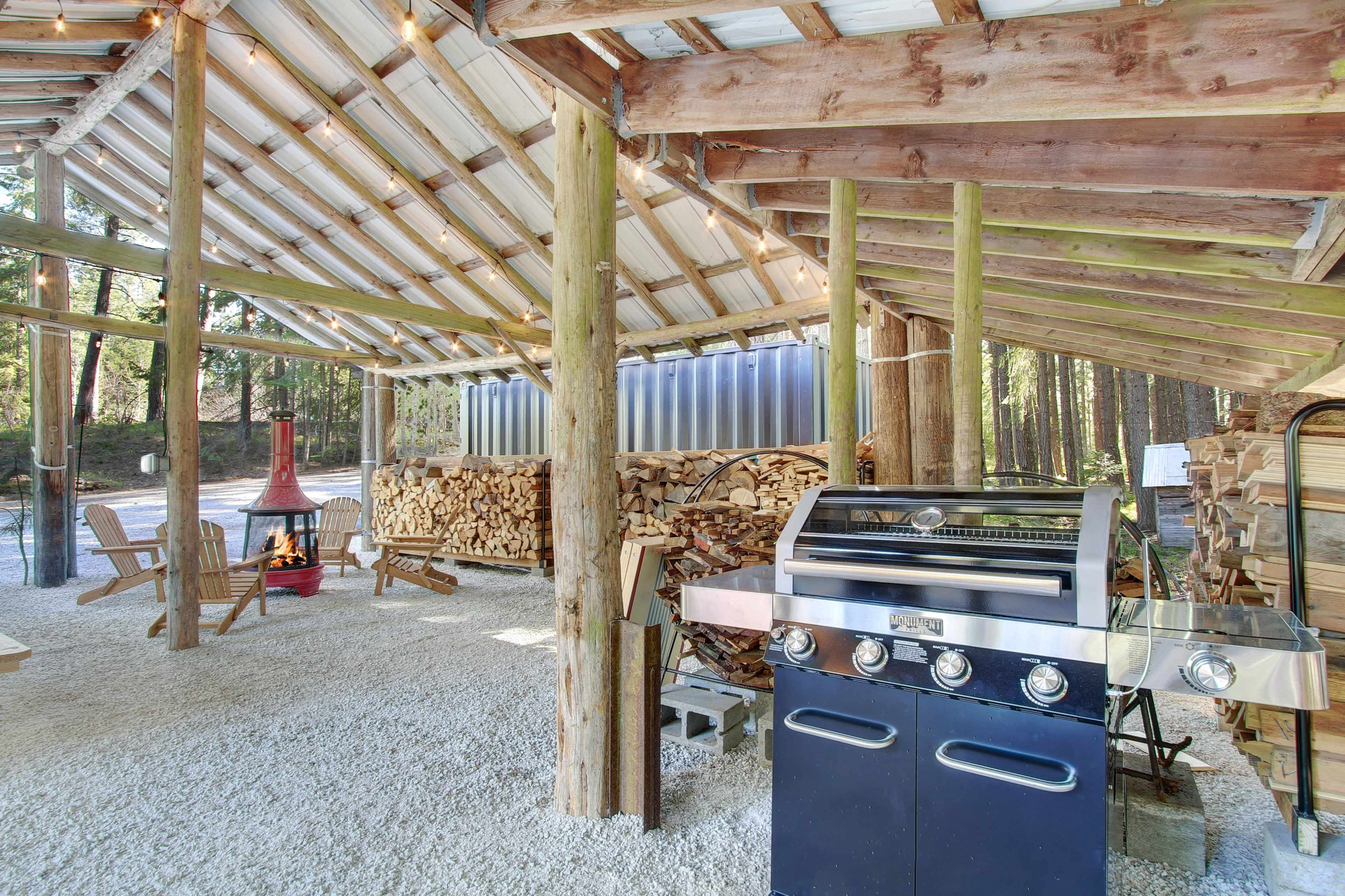 The image shows an outdoor cooking area with a grill, a red fireplace, and stacks of firewood under a roof supported by wooden beams.