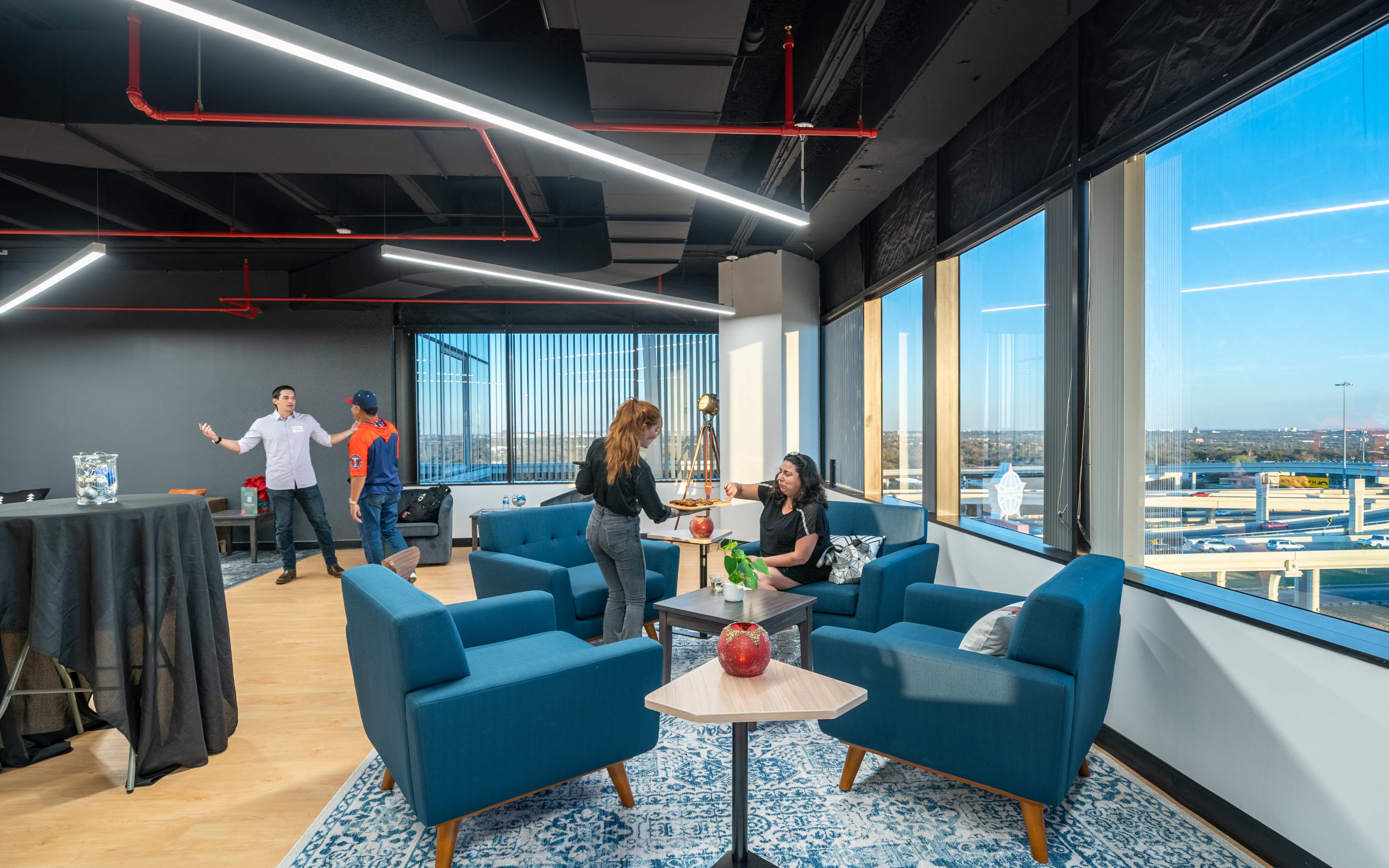 A modern office lounge area with blue chairs, a coffee table, and large windows overlooking a cityscape, where two people are interacting while another pair stands in the background.
