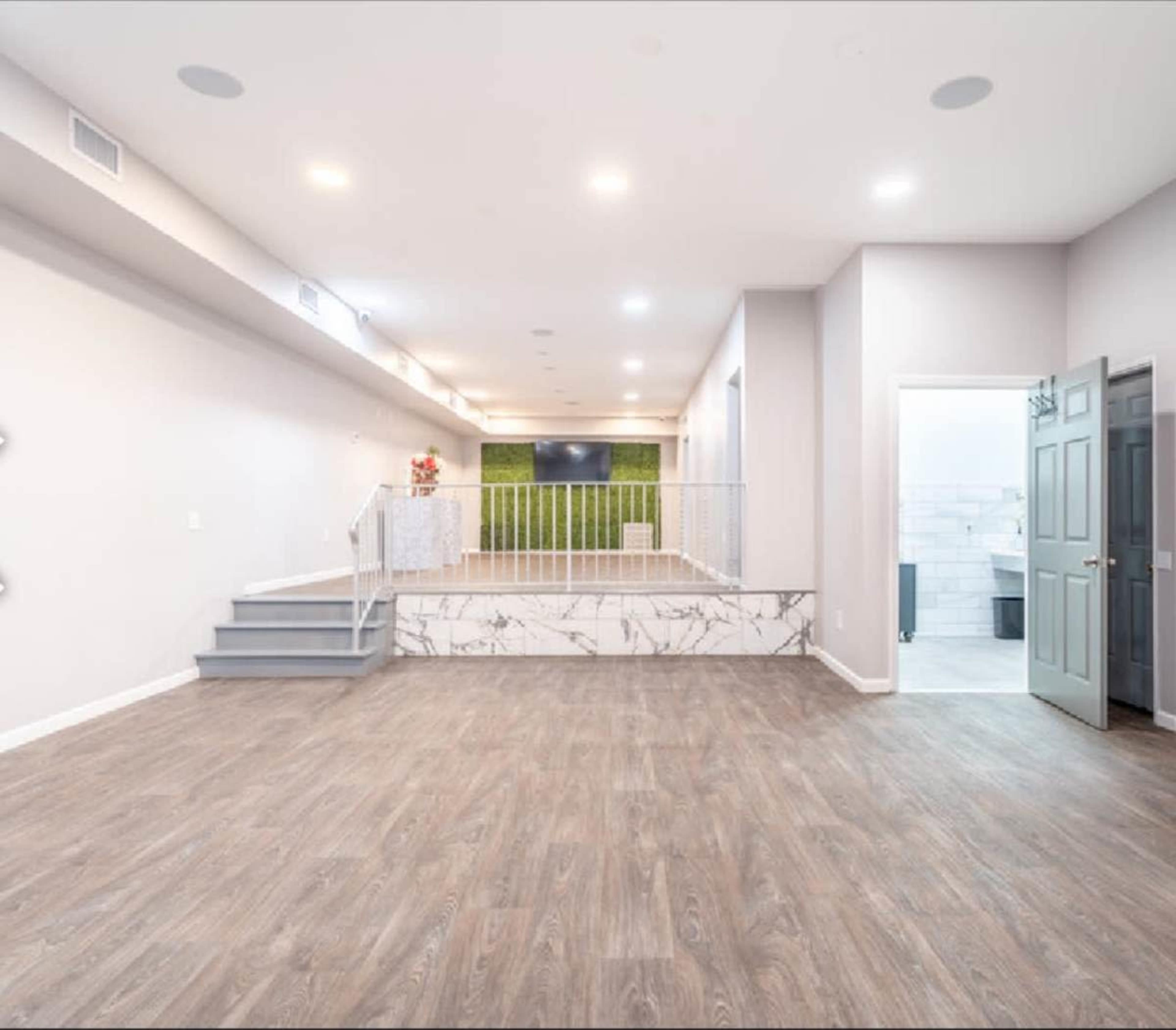 A spacious, modern interior living area with a staircase, a large television mounted on the wall, and an open doorway leading to another room.