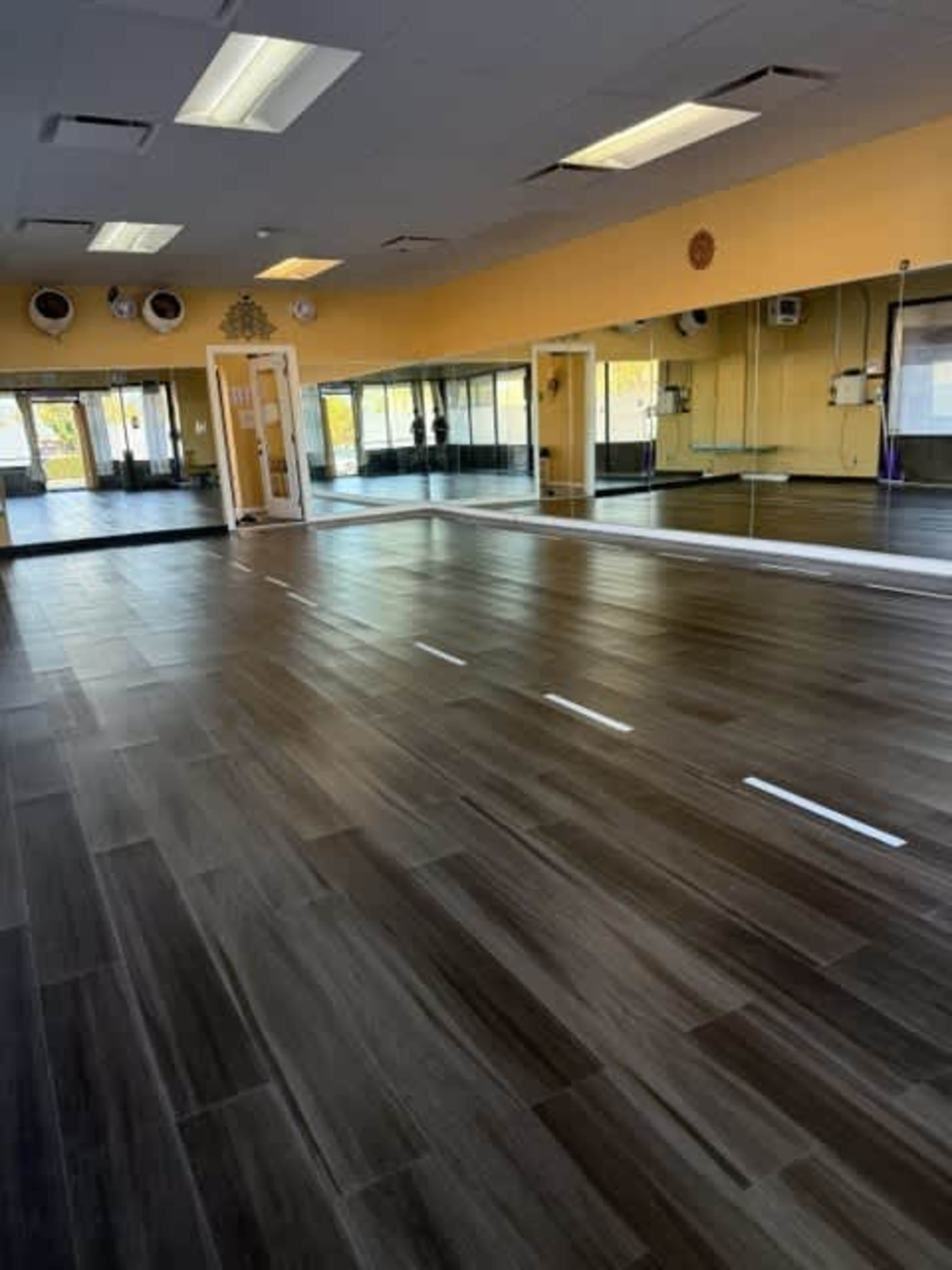 The image shows a spacious room with wooden flooring, large mirrors on one wall, and sunlight streaming in from windows.