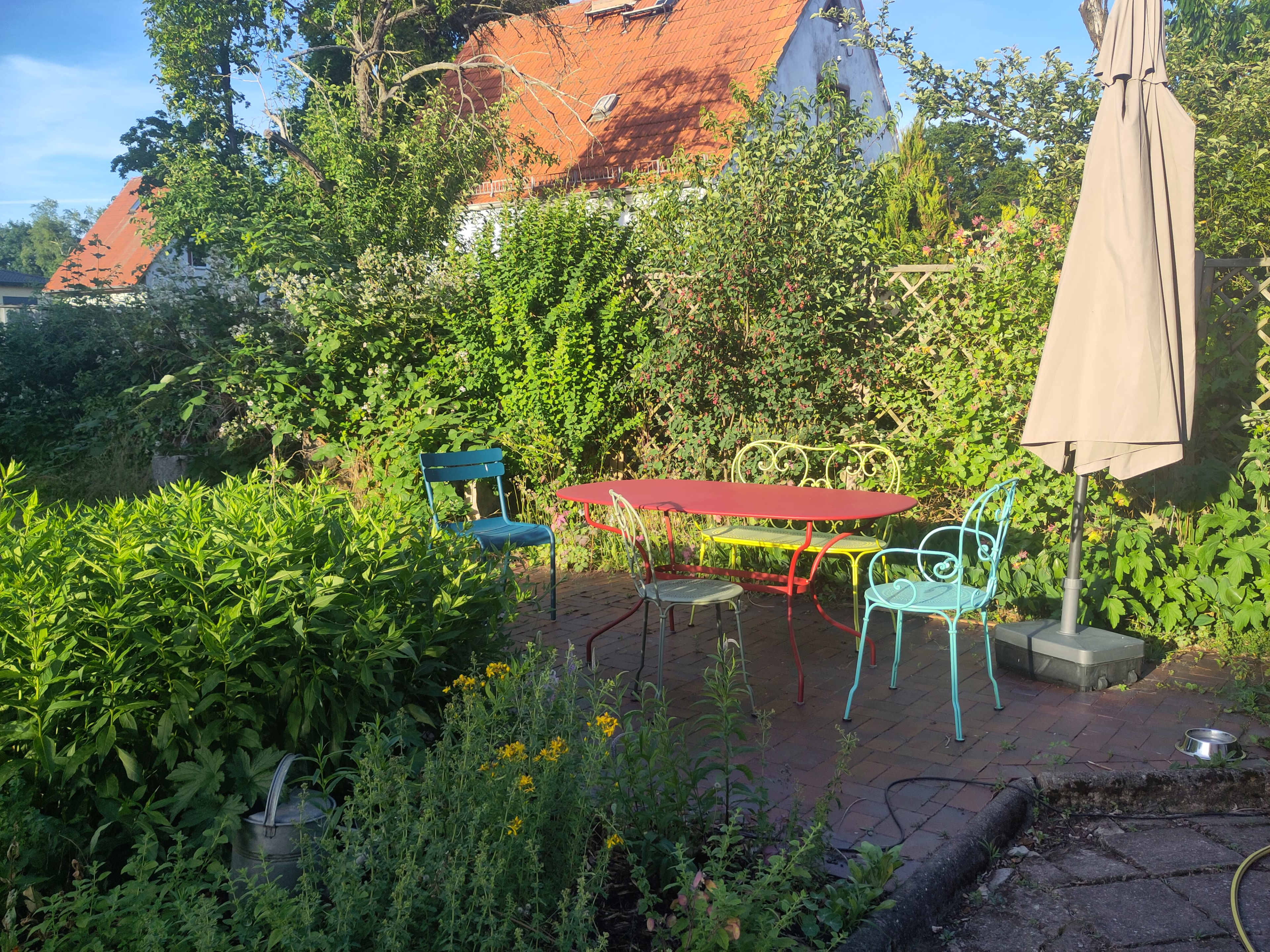 The image shows a colorful outdoor seating area with a red table and four chairs, surrounded by lush green plants and trees, with a patio umbrella.
