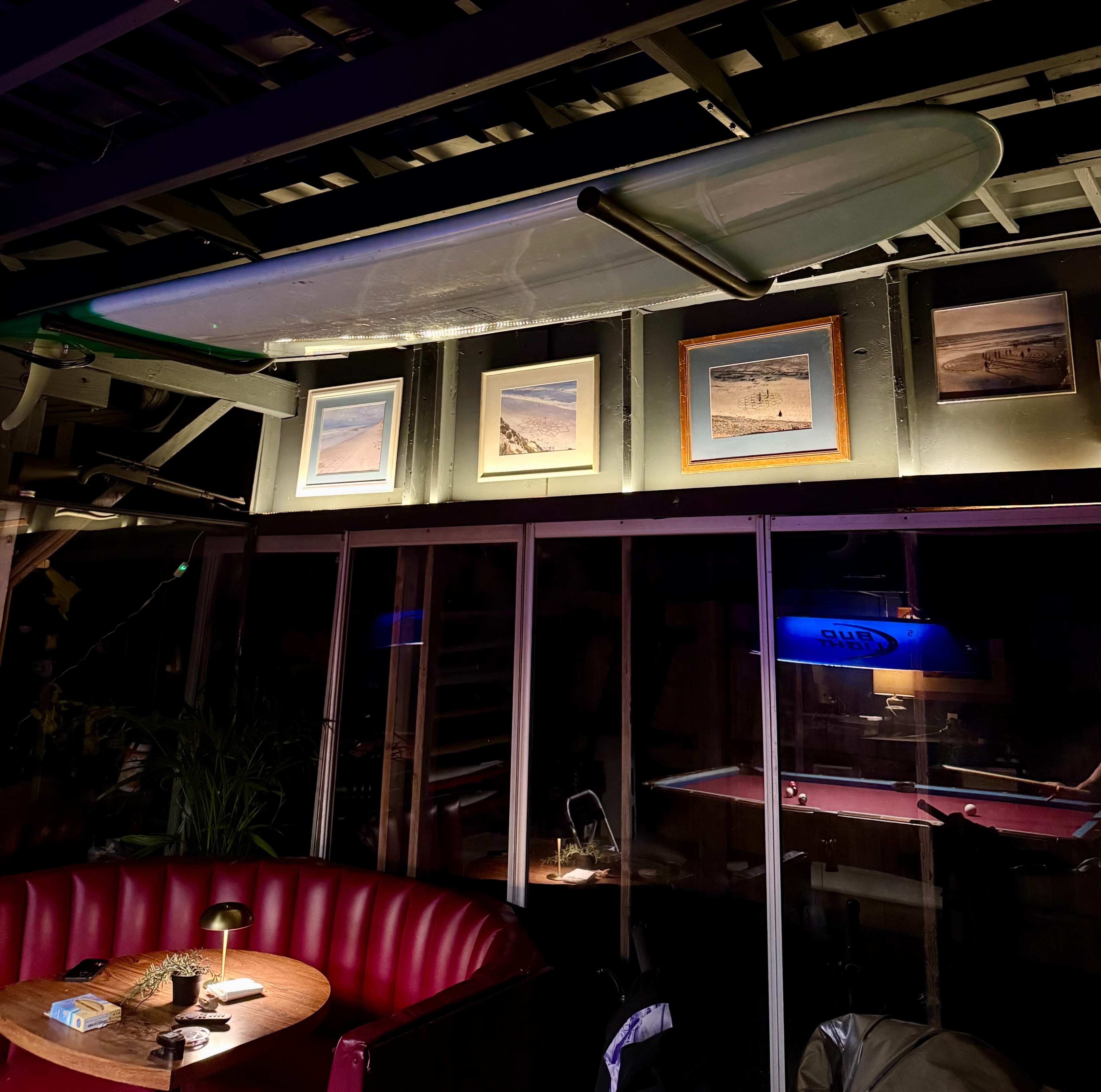 The image shows a bar or lounge area with a surfboard hanging overhead and framed beach photographs mounted on the wall.