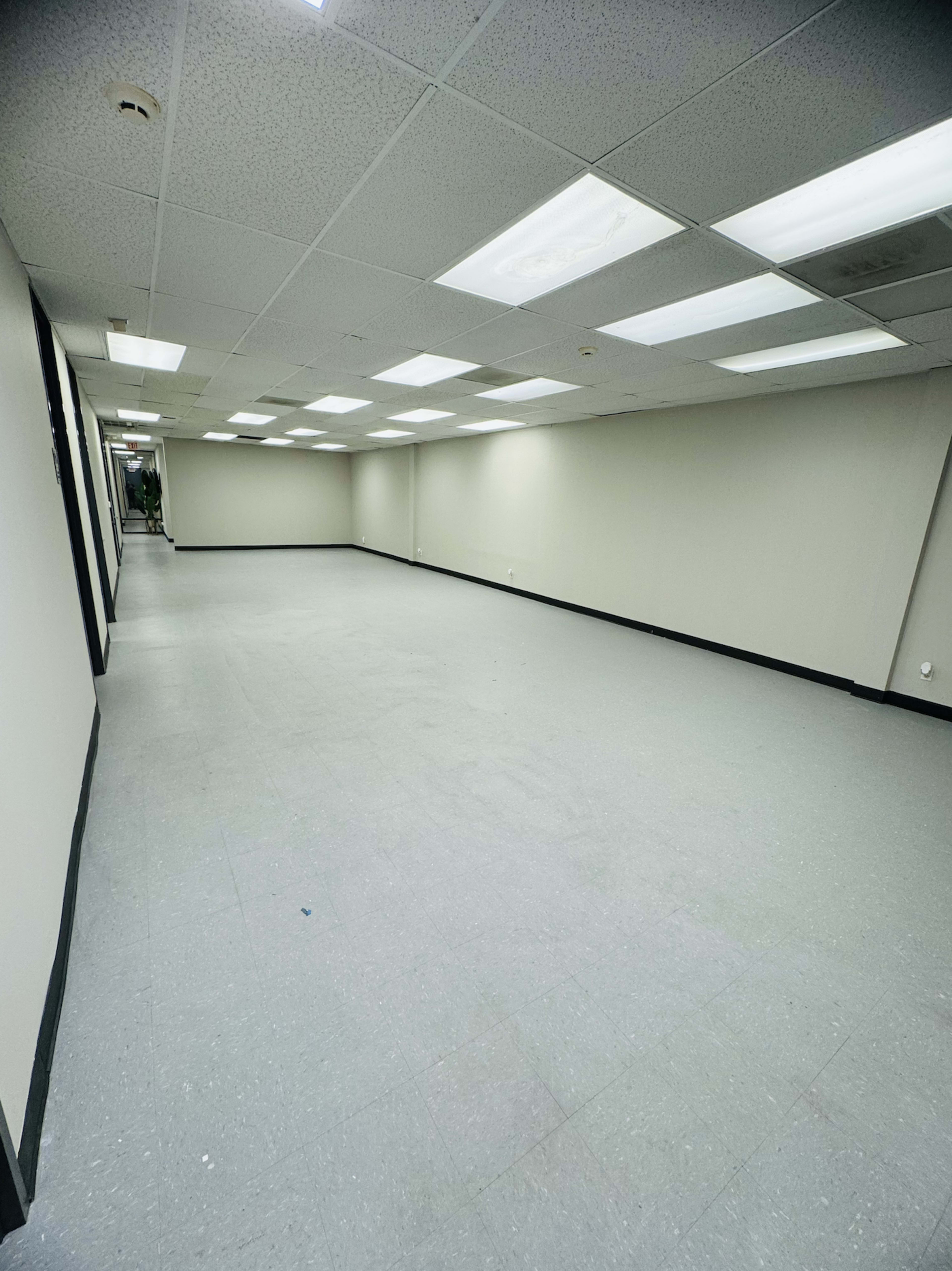The image shows a large, empty room with gray flooring and fluorescent lighting, surrounded by white walls.