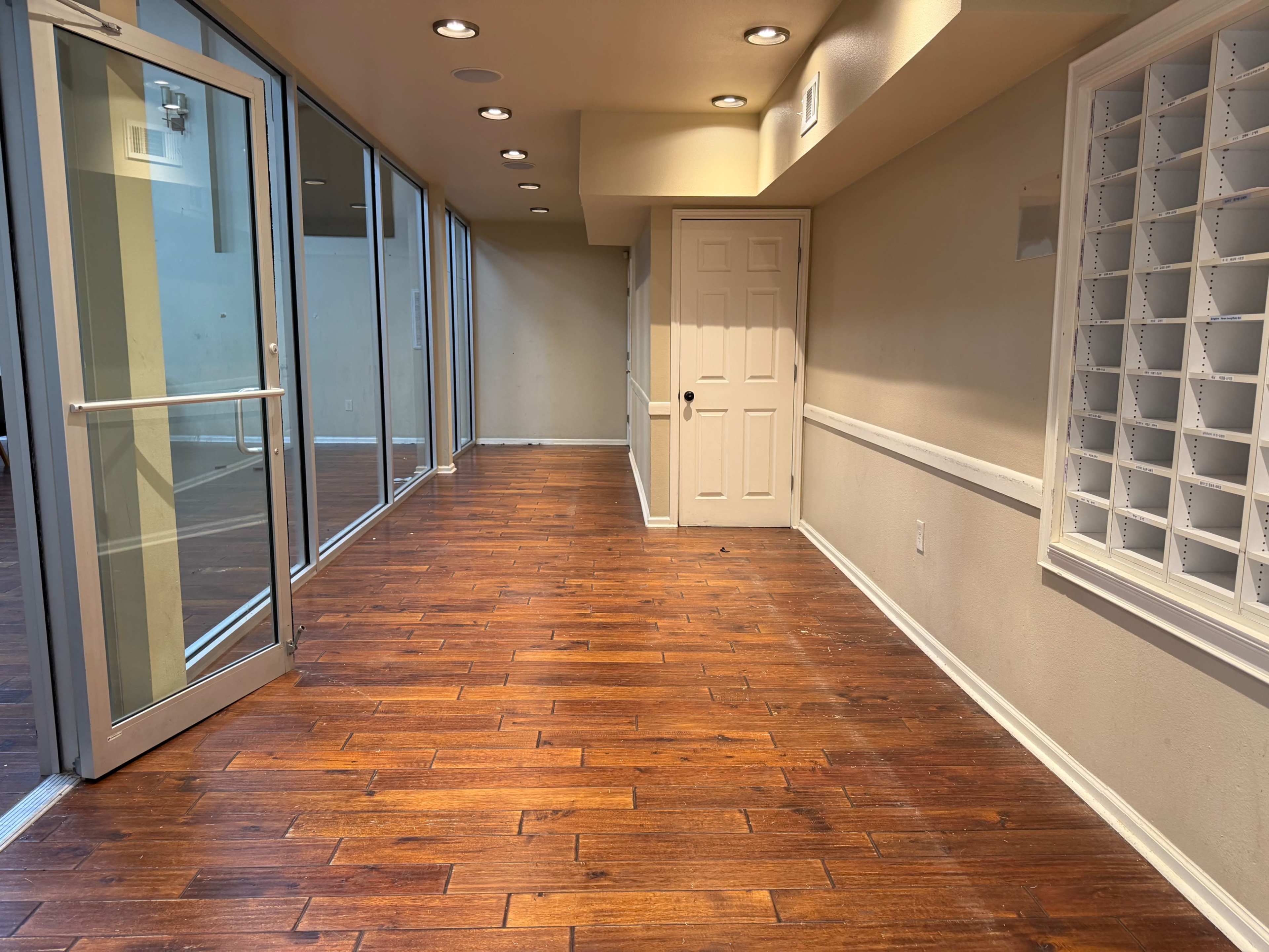 The image shows a spacious interior room with hardwood flooring, large glass doors on one side, and a door leading to another area, along with a wall-mounted mail slot fixture.