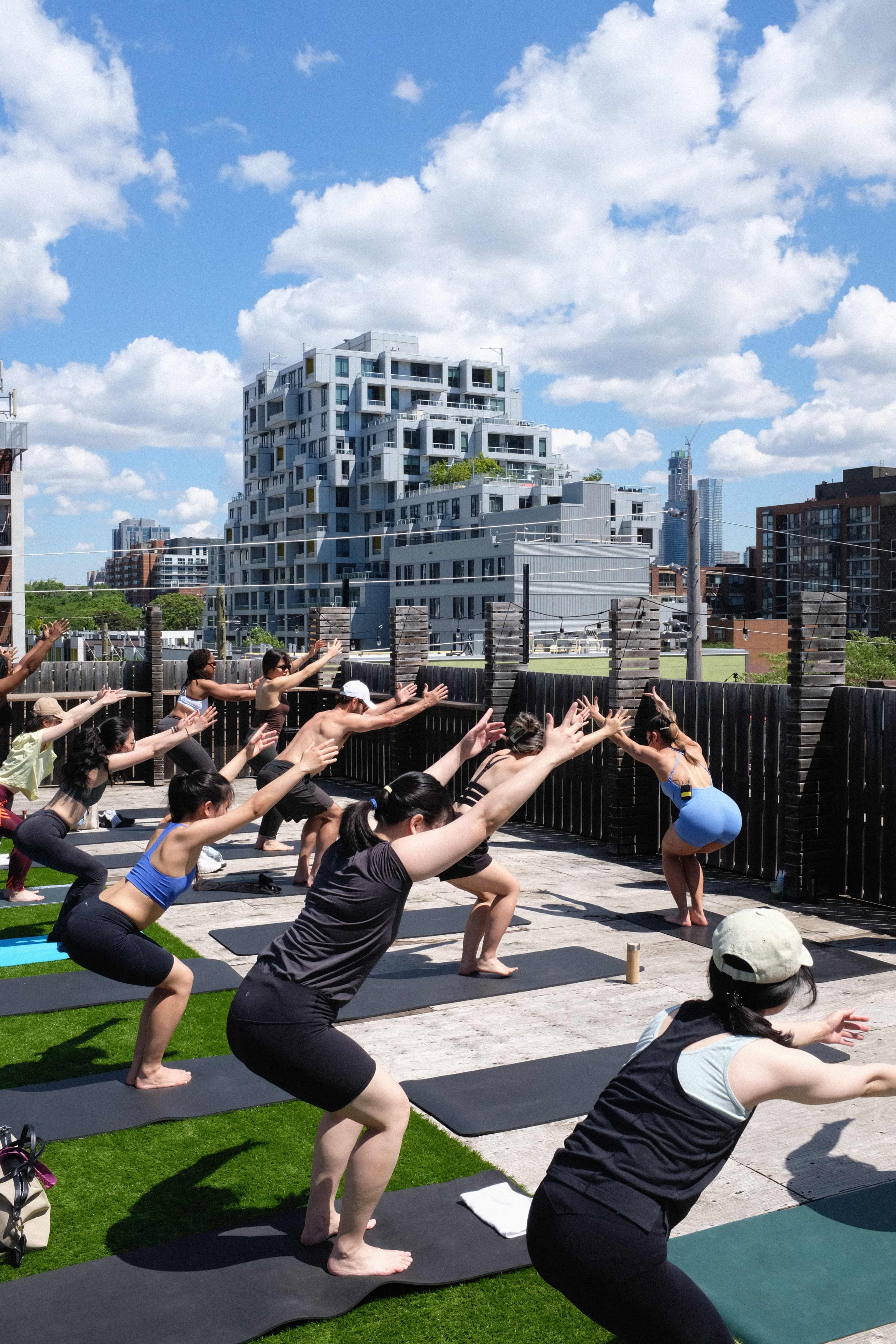 A group of people is practicing yoga in a downward squat position on a rooftop with a city skyline in the background.